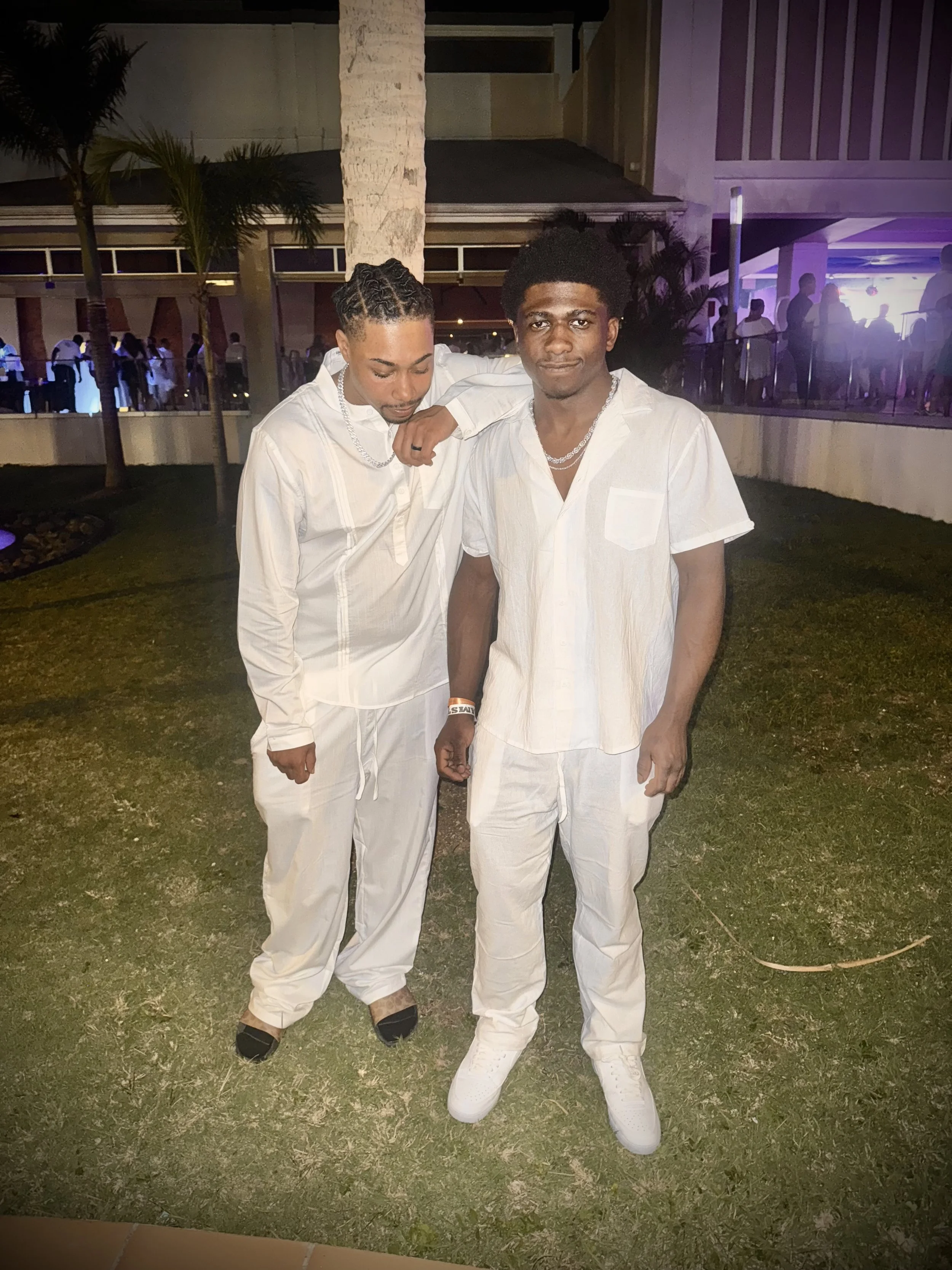 Two young men dressed in white standing outdoors at night, with a background of trees, a building, and other people at a social event.