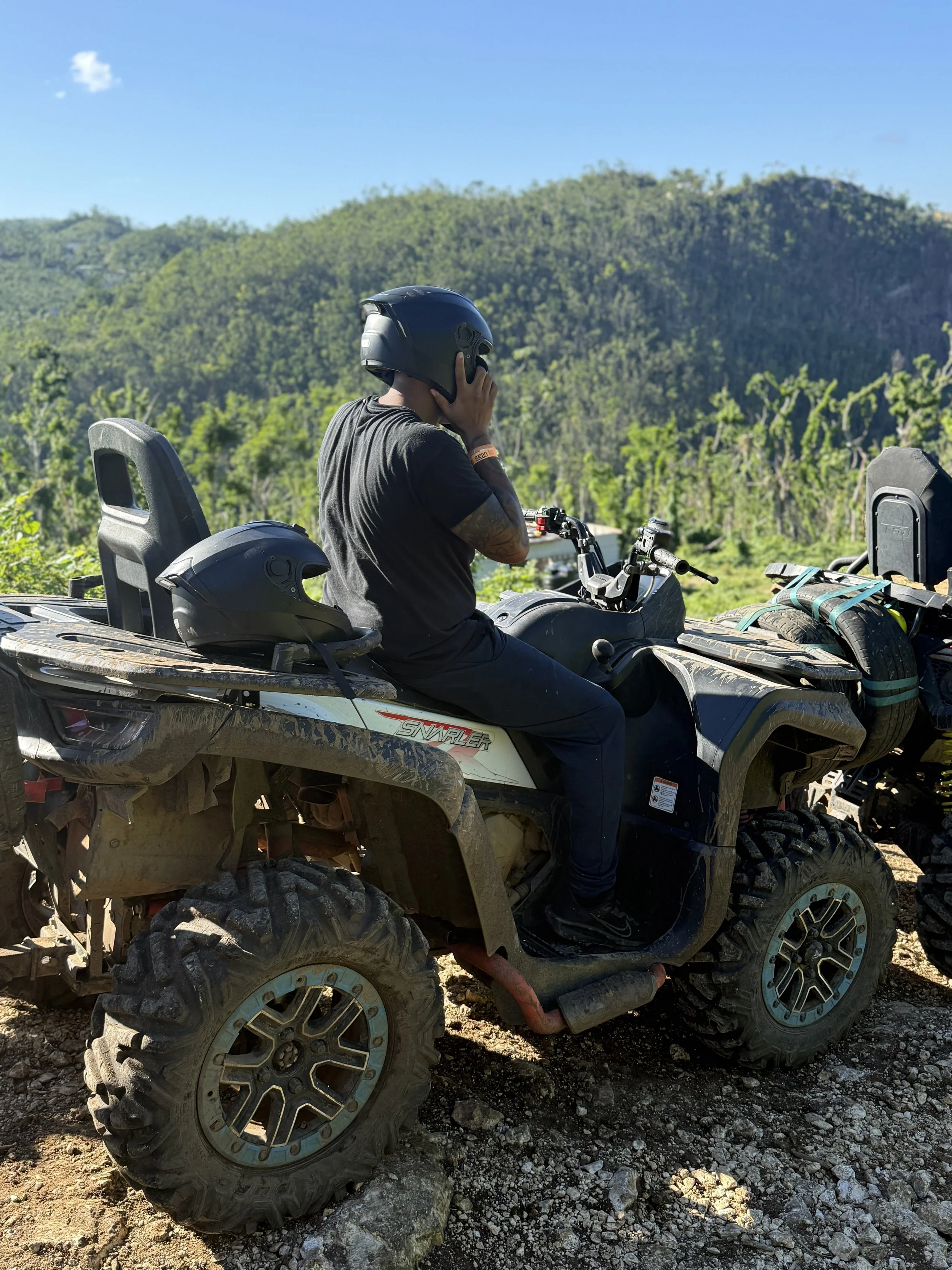 Person sitting on a side-by-side off-road vehicle with a black helmet on the seat, surrounded by a rocky terrain and green hills in the background, under a blue sky.