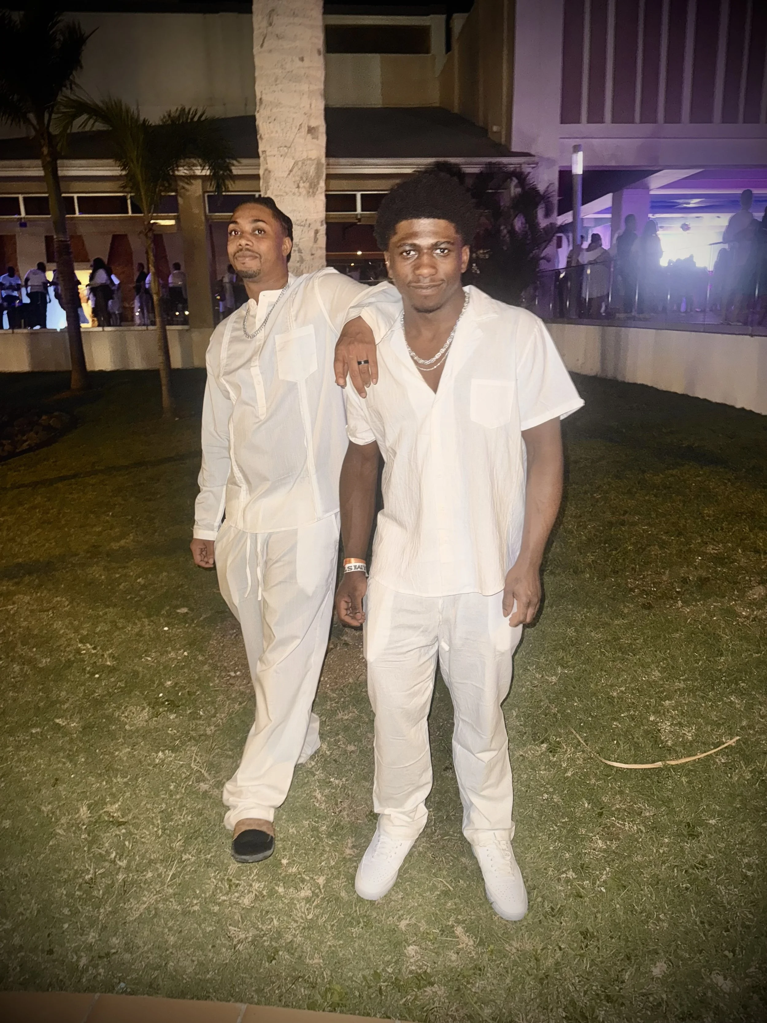 Two men posing together outside at night, both wearing white clothing. One man has his arm around the other's shoulder. The background appears to be a party or gathering with other people, some with illuminated screens.