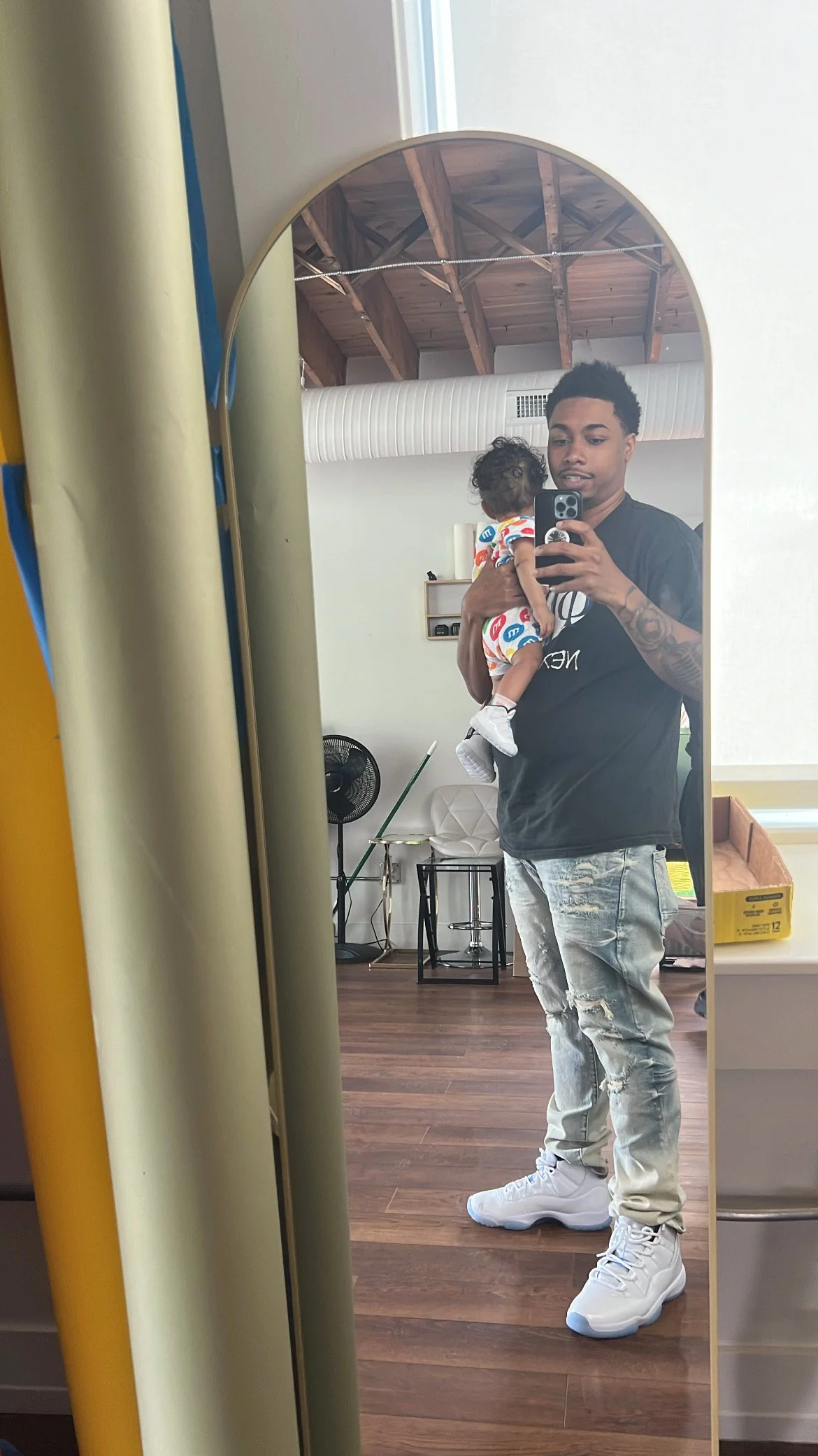 A man taking a selfie in a mirror while holding a small child. The man is wearing a black t-shirt and ripped jeans, and the child is wearing a colorful outfit with a pattern. The room has a wooden ceiling, a white chair, a fan, and various objects on