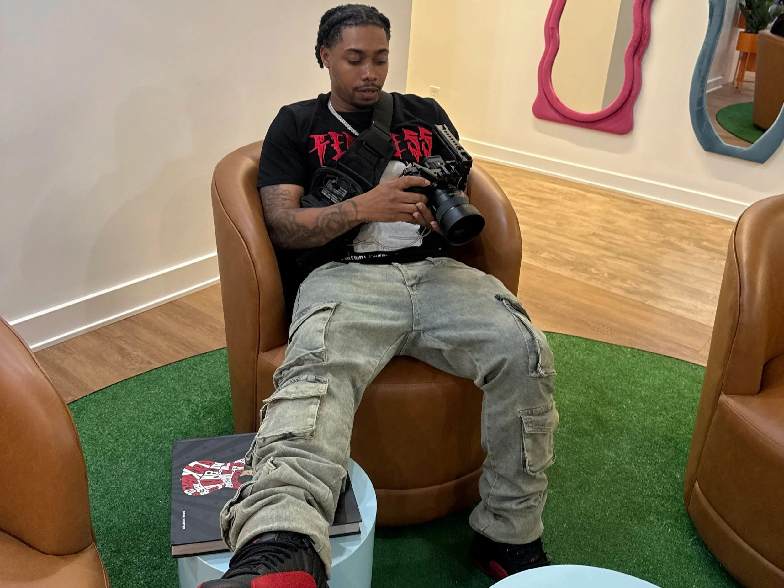 A young man with dreadlocks sitting in a brown leather chair, looking at a camera he is holding. He is wearing a black T-shirt with red text, light-colored cargo shorts, and black sneakers. He is seated in a room with light-colored walls, colorful irregularly shaped mirrors on the walls, wooden flooring, a green carpet, and a small table with a book underneath him.