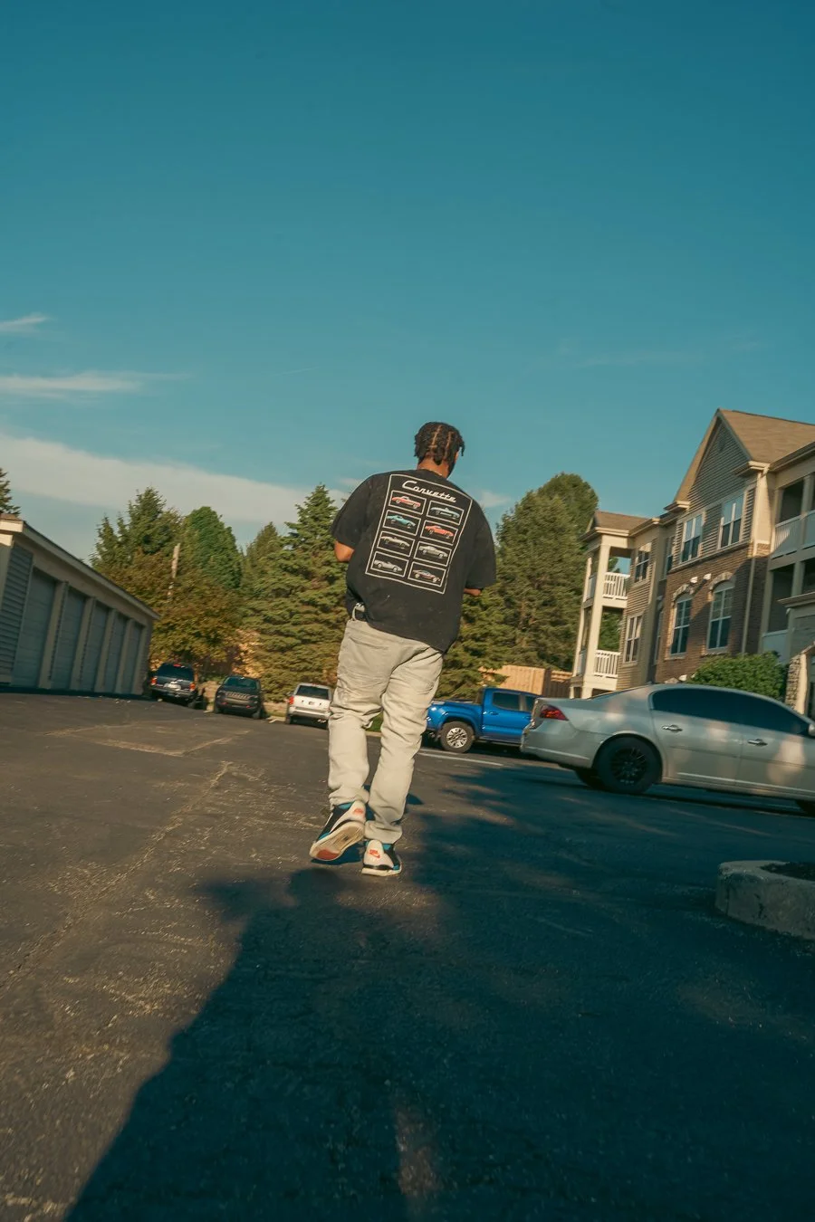 A person walking in a parking lot during daylight, wearing a black T-shirt with car illustrations on the back and light-colored pants, with residential buildings and parked cars in the background.