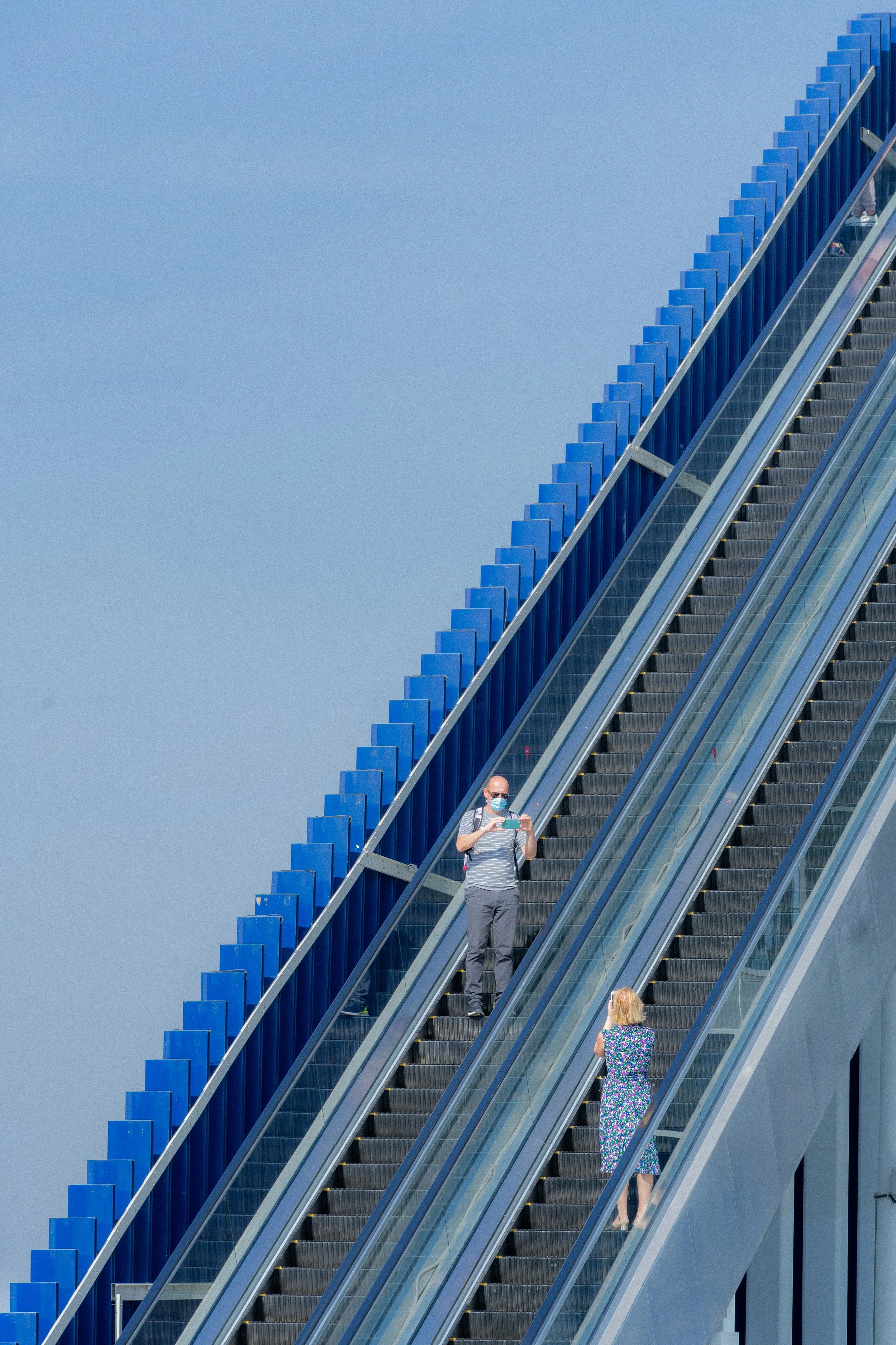 Une personne avec un masque facial, un t-shirt rayé gris et blanc, et un pantalon gris, tenant un téléphone portable, descend un escalator extérieur avec un mur en béton et un ciel bleu clair en arrière-plan. Au bas de l'escalator, une femme avec une
