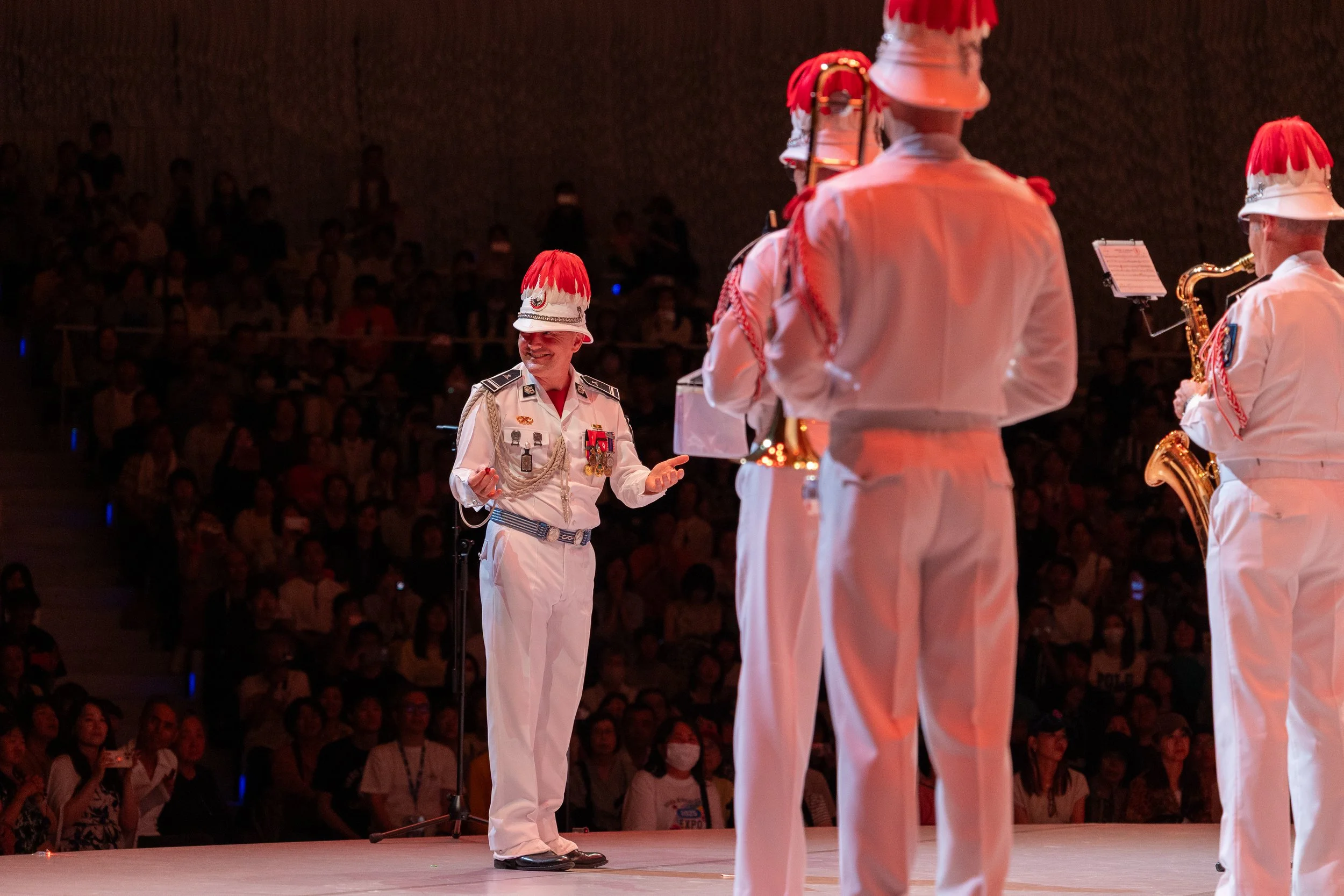Musiciens militaires en uniforme blanc avec chapeaux à plumes rouges jouent de la musique lors d'une cérémonie sur scène, avec un public en arrière-plan.