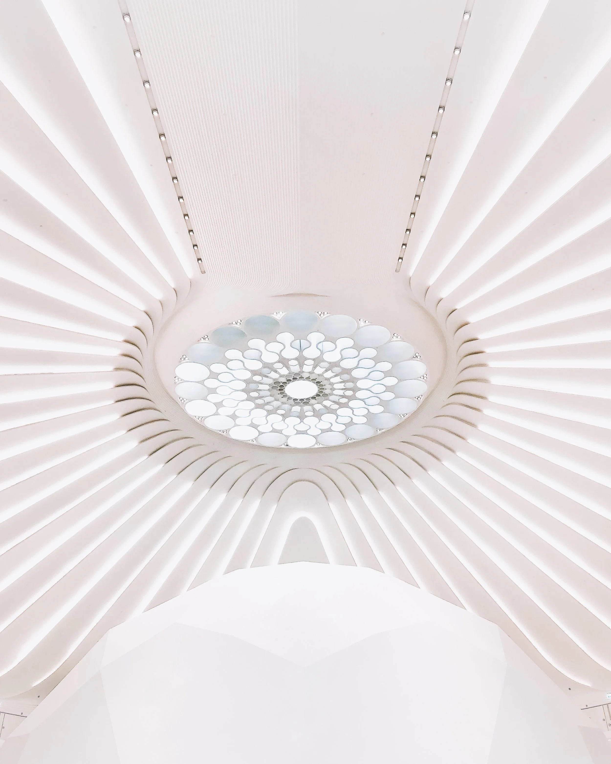 Une vue en plongée d'un plafond moderne avec des lignes courbes blanches éclairées, des structures ovales en verre ou en cristal laissant passer la lumière, création architecturale élégante et lumineuse.