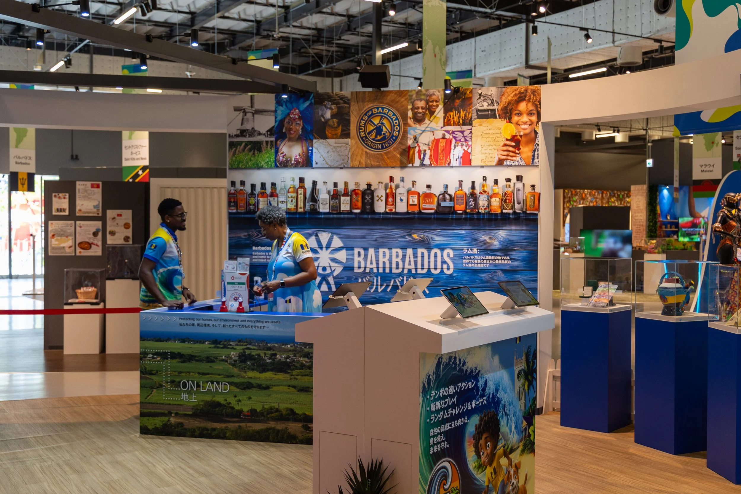 Stand d'information de l'île de la Barbade avec des affiches colorées, une étagère de bouteilles d'alcool, deux personnes en conversation, et des images de la culture et du paysage de la Barbade.