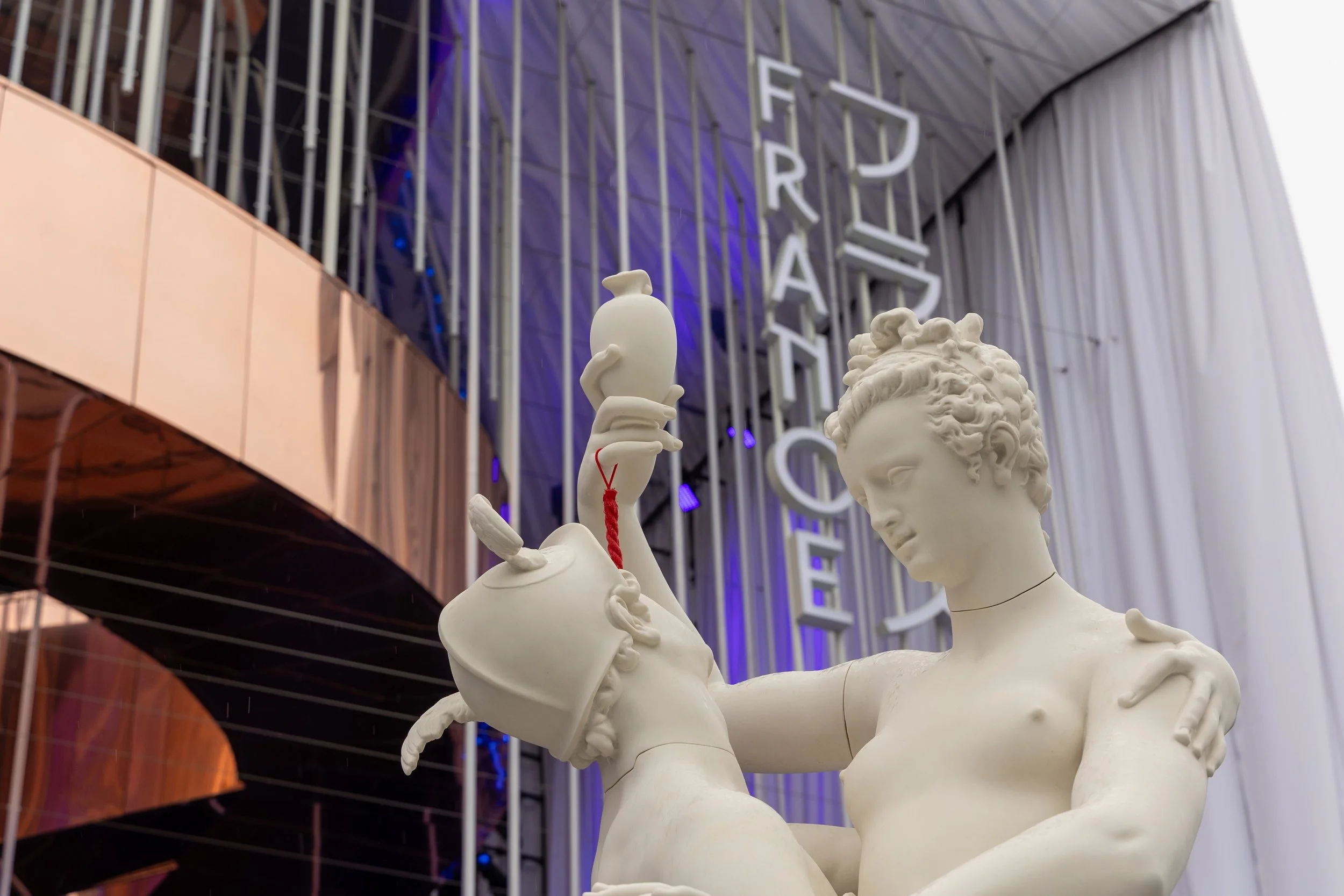 Sculpture blanche représentant une femme avec une couronne de fleurs dans les cheveux, tenant une lampe dans la main, située devant un bâtiment avec un panneau lumineux vertical."