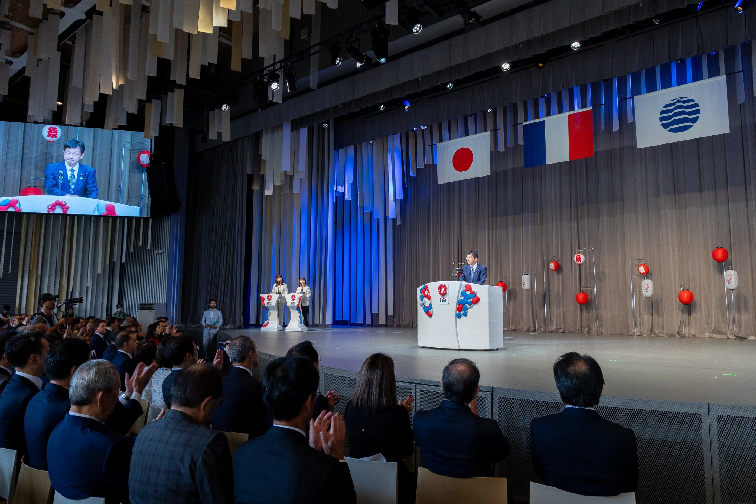 Une grande scène avec des personnes en train de faire un discours ou une présentation, décorée avec des drapeaux internationaux dont ceux du Japon, de la France, et un drapeau avec des rayures bleues et blanches. Il y a un public assis regardant la s