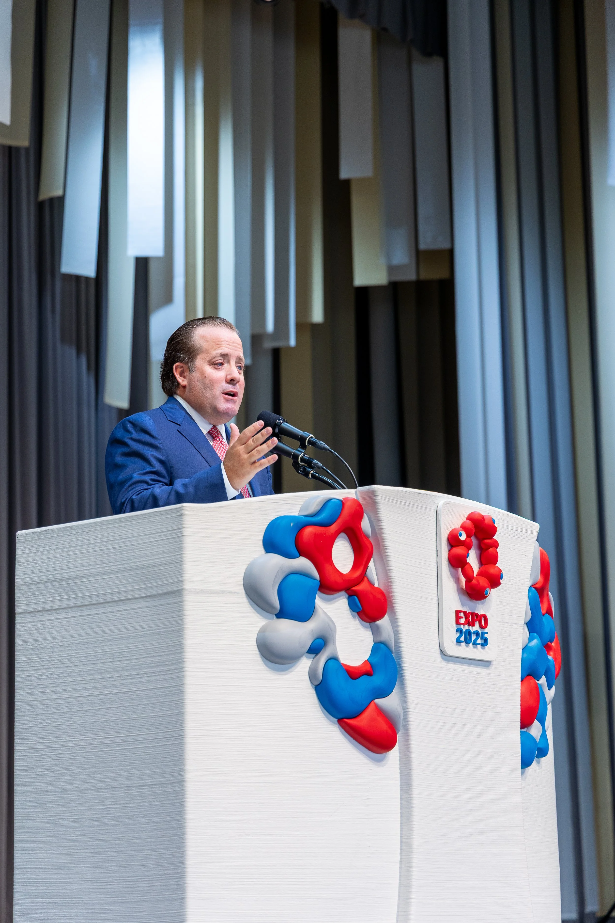 Un homme en costume blue parle à un micro lors d'un événement, avec un décor de formes géométriques colorées en rouge, blanc, et blue, portant le logo 'Expo 2025'.