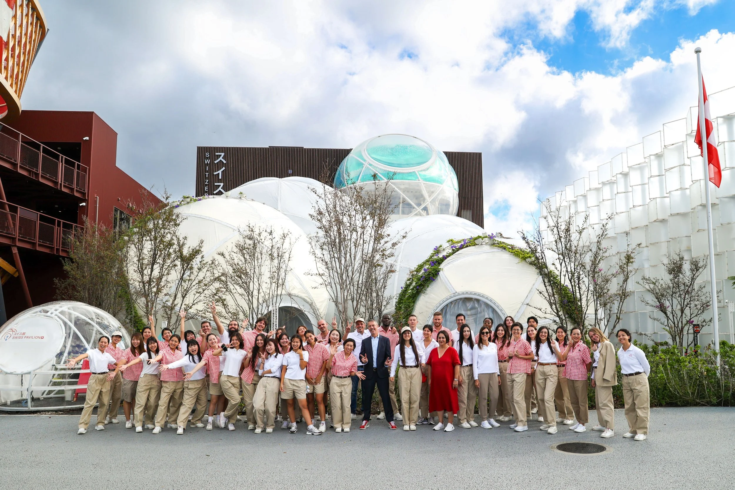 Groupe de personnes souriantes posant devant le pavillon suisse lors d'une sortie touristique, avec des structures géodésiques en plastique transparent, un bâtiment moderne avec enseigne en japonais et anglais, et un ciel partiellement nuageux.