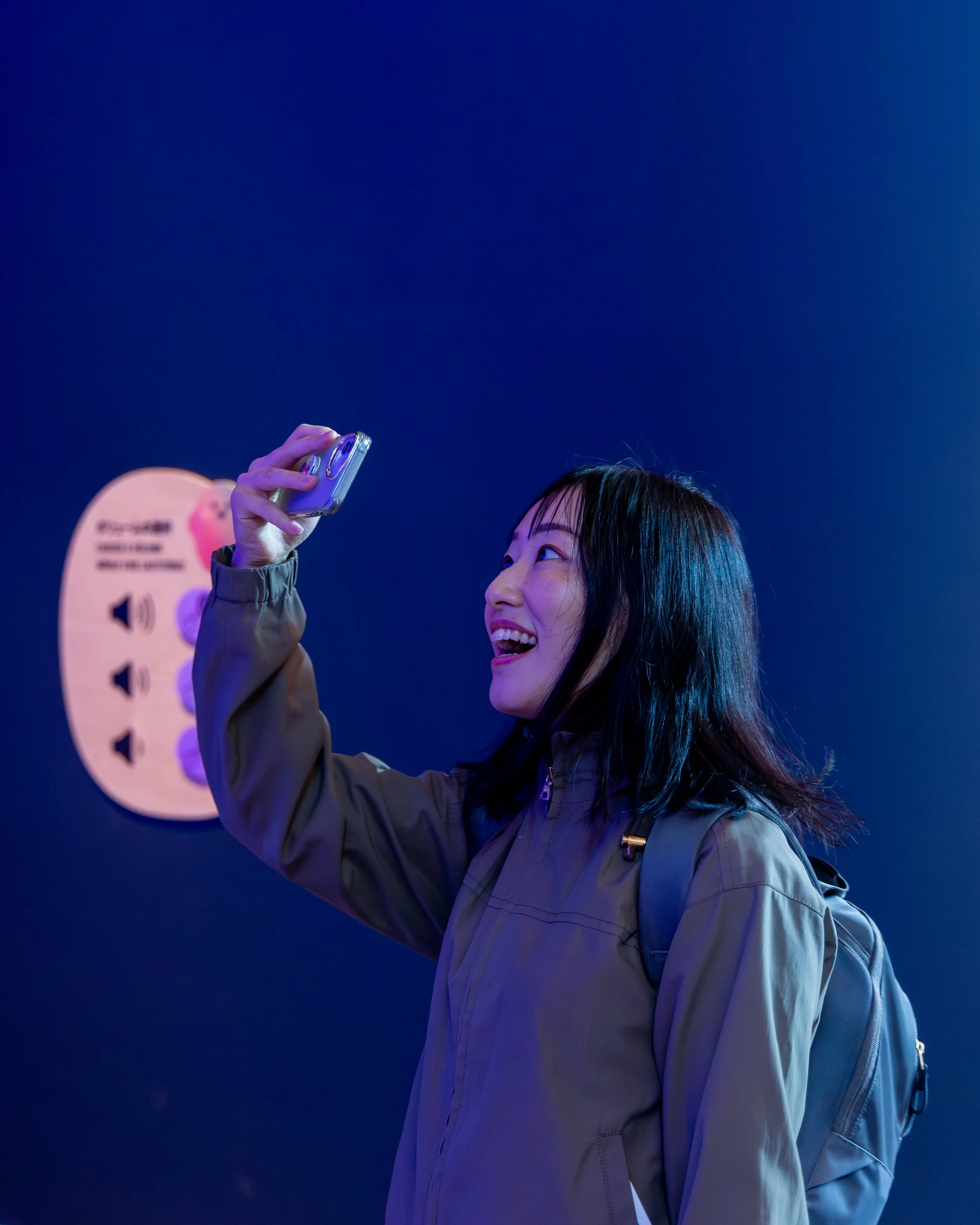 Une jeune femme souriante portant un manteau beige et un sac à dos violet, tenant un téléphone portable pour prendre un selfie, avec un fond bleu foncé et un panneau en arrière-plan.