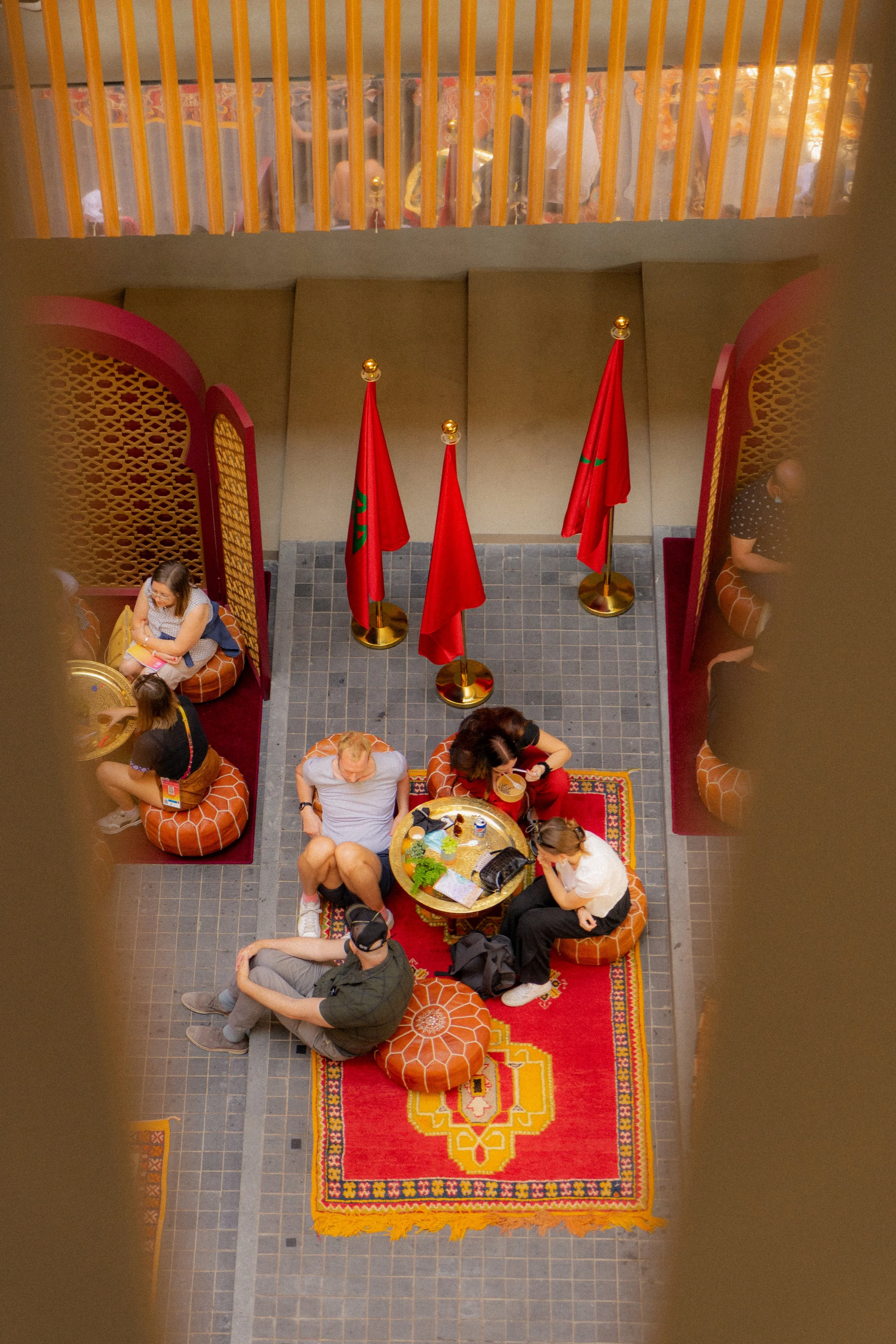 Vue intérieure d'un hall avec des personnes assises autour d'une table, des drapeaux rouges avec un emblème vert, des paravents rouges et une moquette rouge avec un motif coloré.