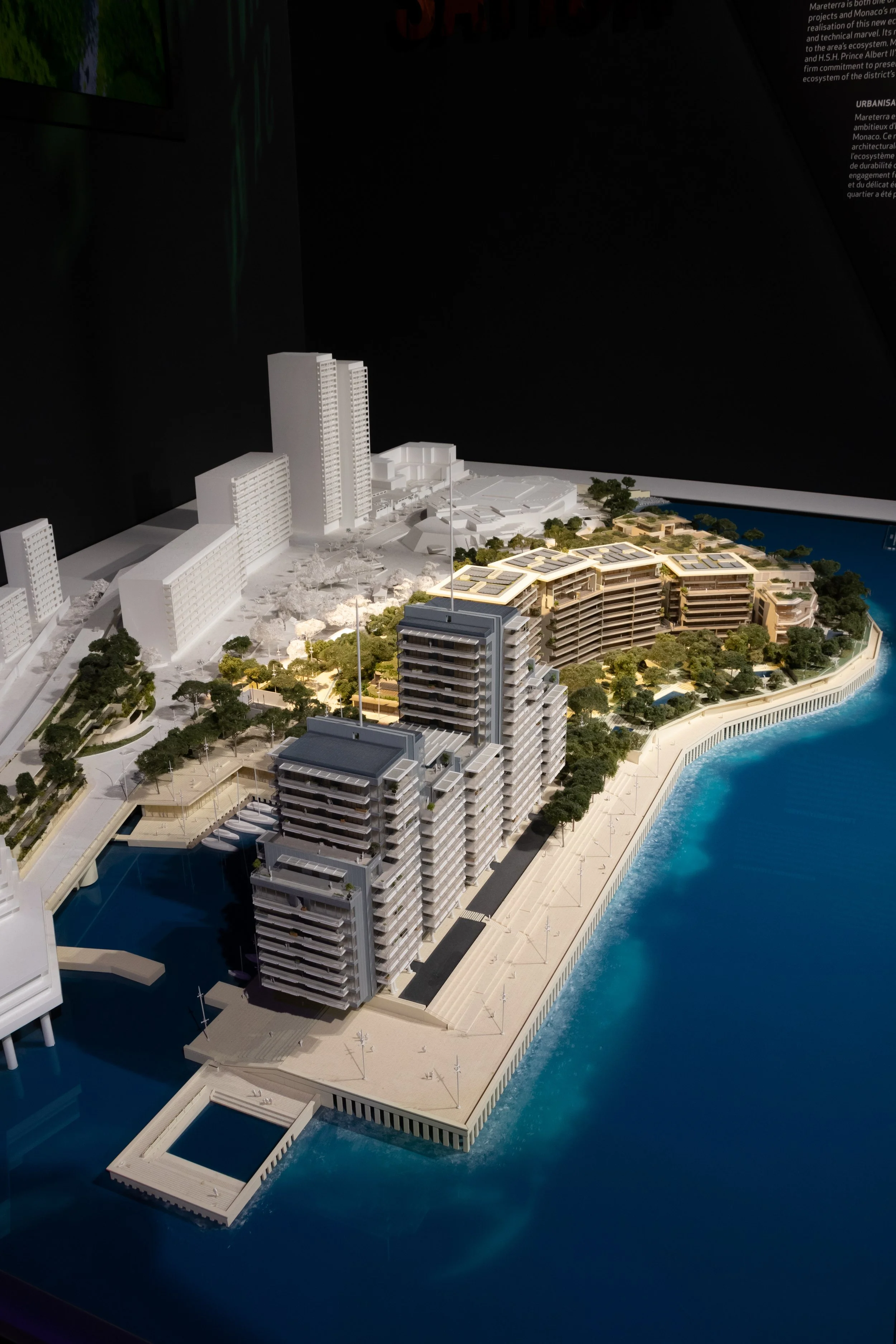 Modèle réduit d'un complexe immobilier sur un plancher flottant au bord de l'eau, avec plusieurs bâtiments modernes, hautes tours, espaces verts et vue sur la mer.
