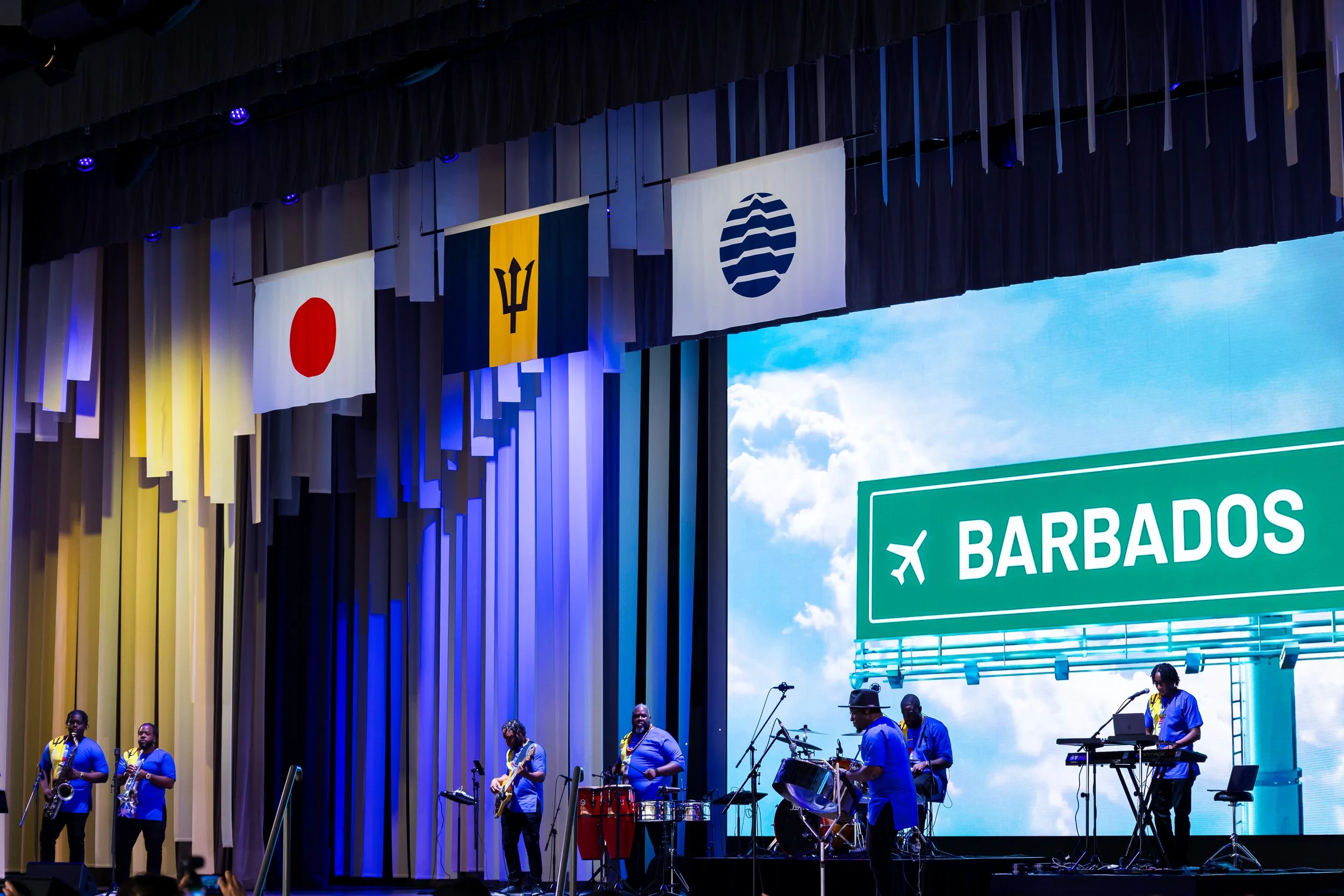 Une scène de concert avec un groupe musical jouant sur scène, des drapeaux de différents pays suspendus au plafond, notamment le Japon, la Barbade, et une autre organisation. Un panneau indique l'arrivée vers Barbados.