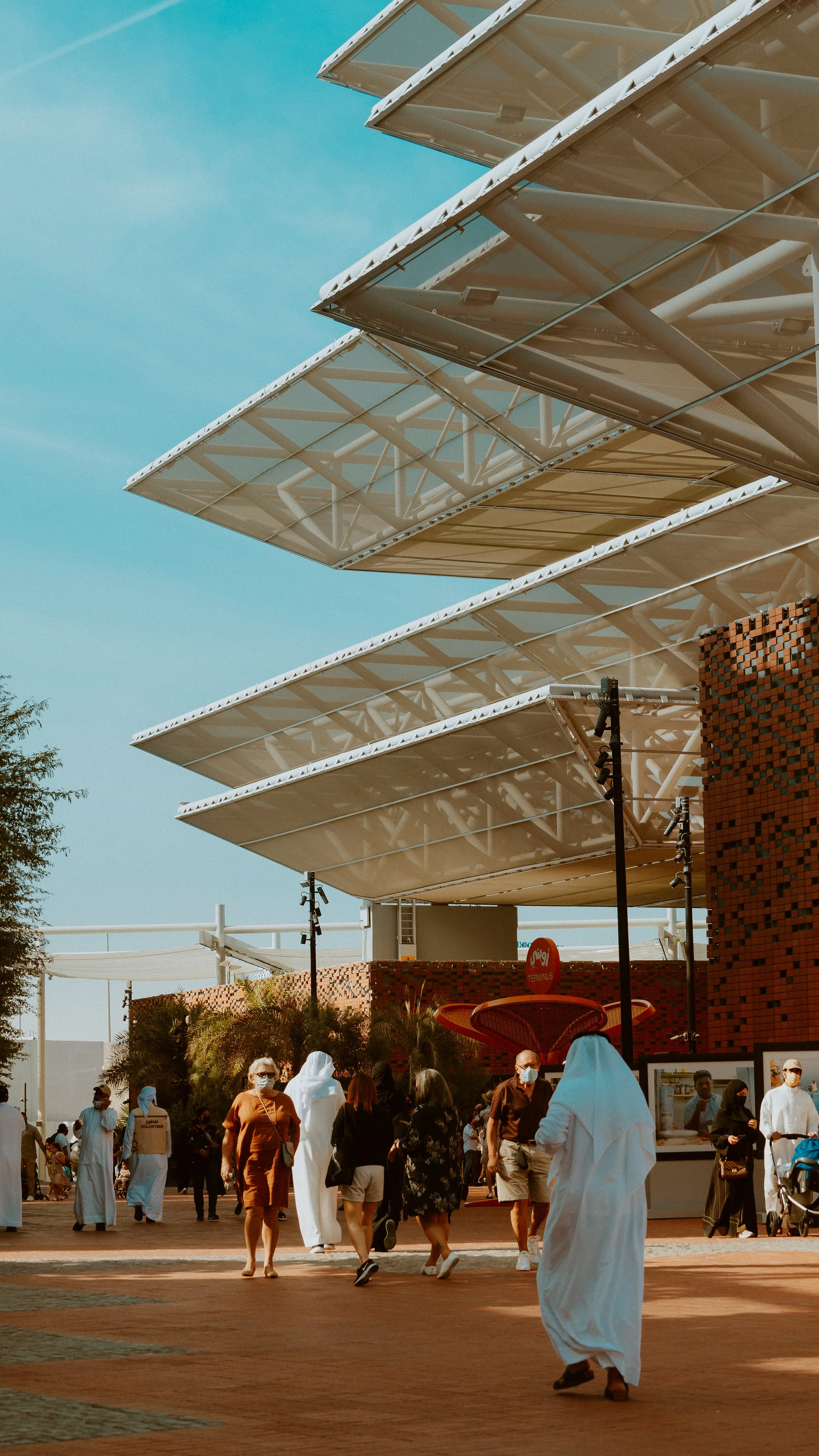 Vue de personnes marchant dans une place publique en plein air, avec un bâtiment moderne en arrière-plan doté d'étages en porte-à-faux et d'une structure métallique visible, sous un ciel bleu clair.
