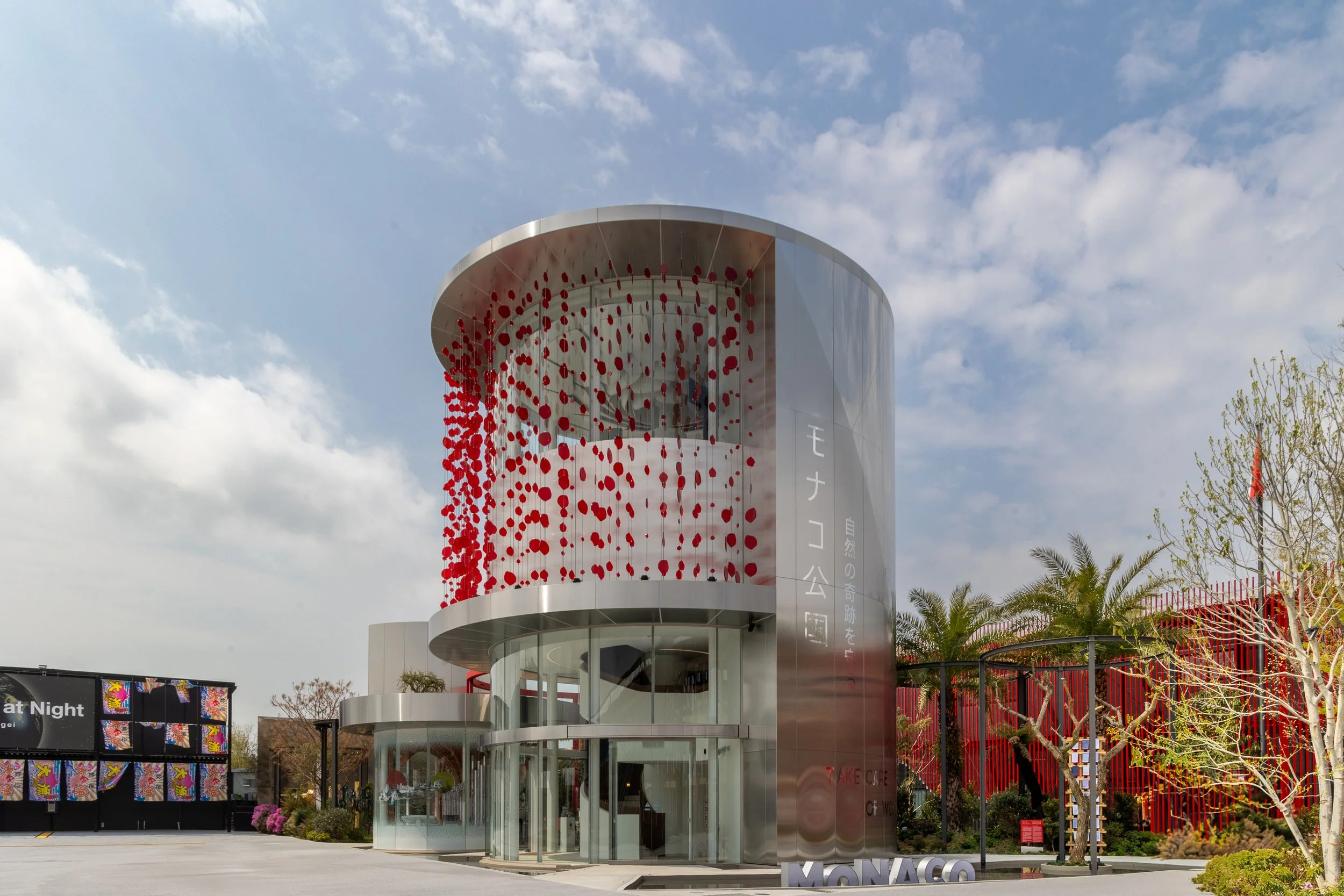 Bâtiment moderne en forme de tour avec façade en métal et des décorations rouge suspendues à l'extérieur, sous un ciel partiellement nuageux, entouré d'arbres et de structures décoratives.