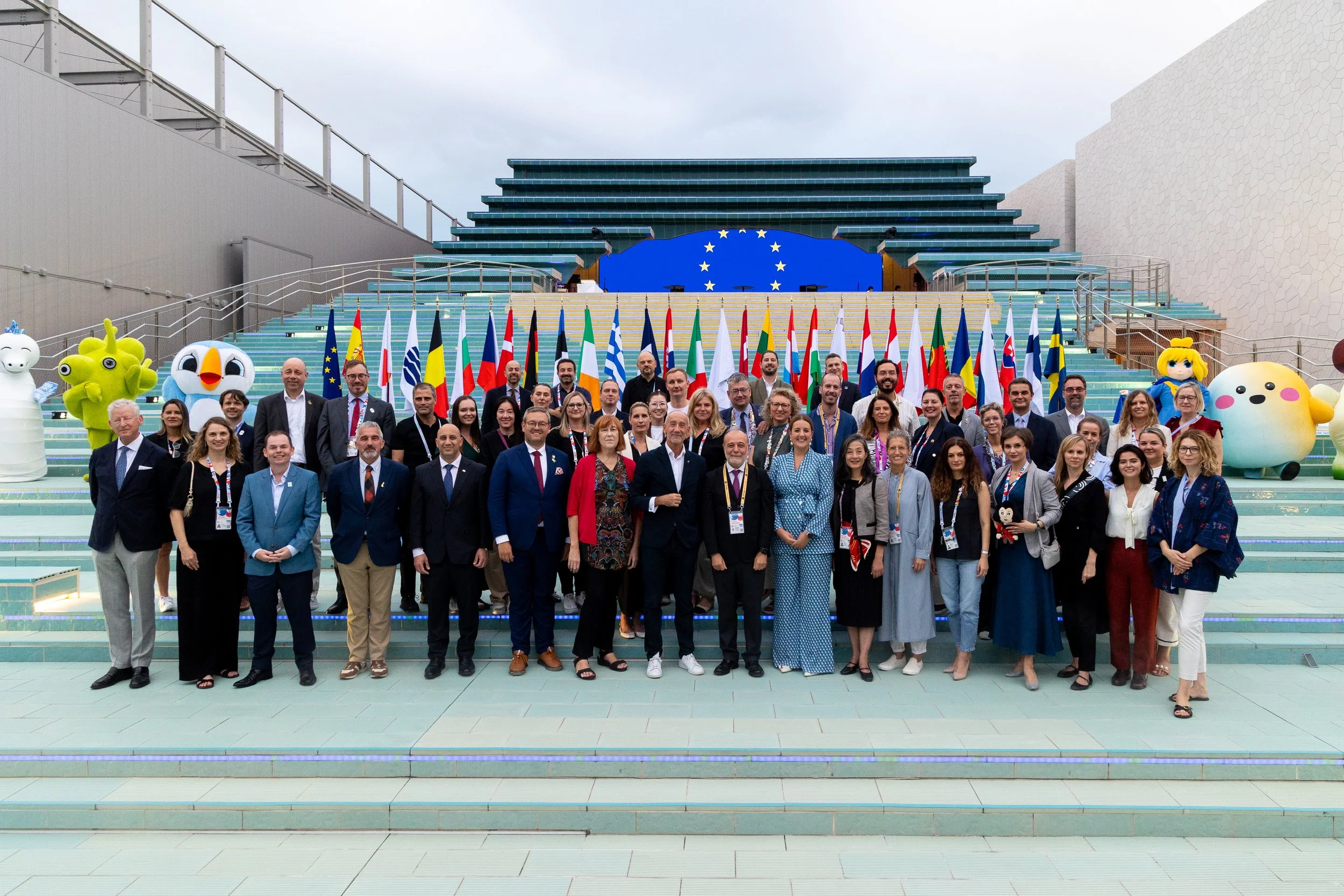 Photo de groupe de personnes devant un bâtiment avec des drapeaux de l'Union européenne, des mascottes et un logo de l'UE, lors d'un événement officiel ou conférence.