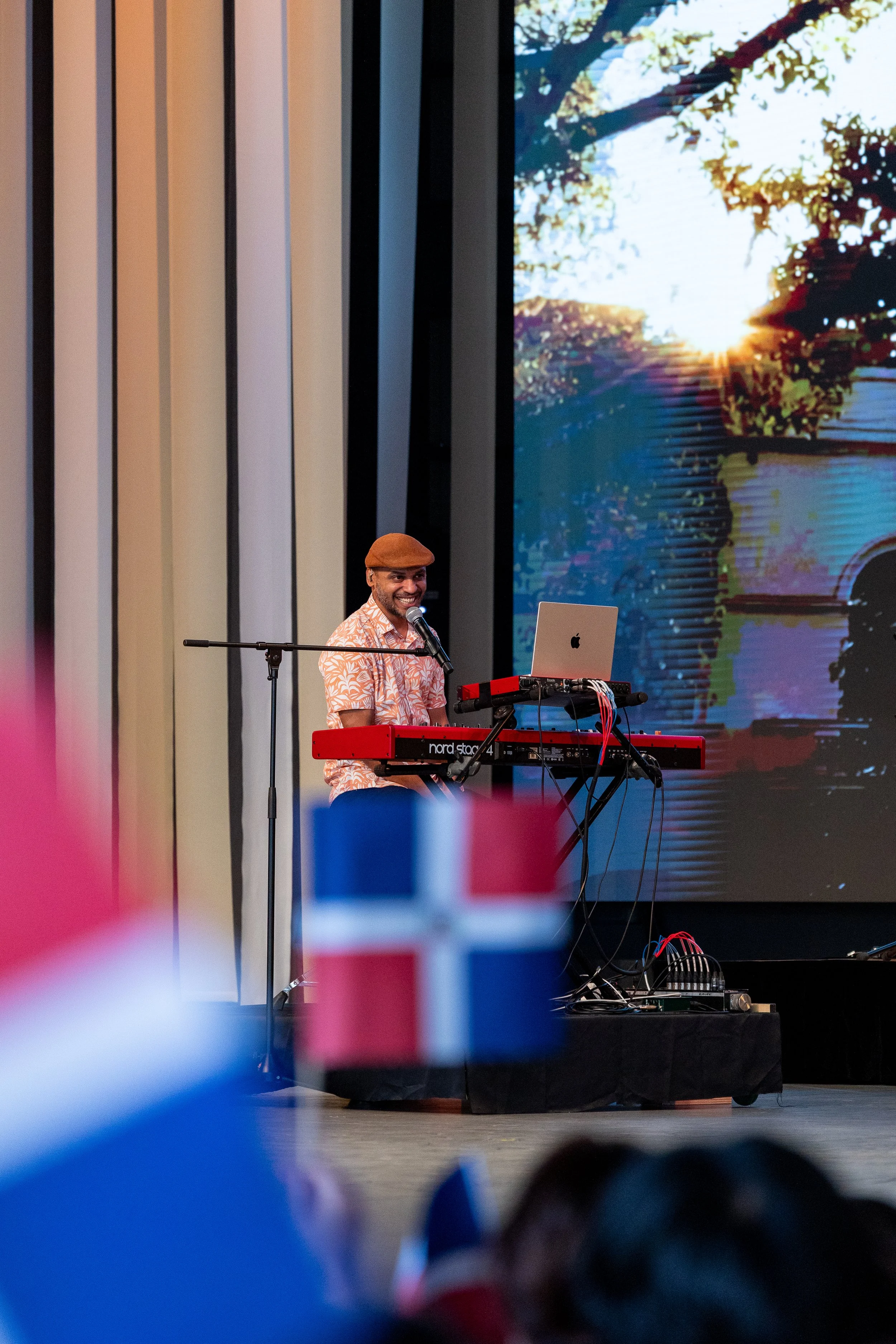 Un homme souriant joue du clavier électrique sur scène, avec un microphone de chaque côté, derrière lui un grand écran affiche une image d'un coucher de soleil à travers des arbres.