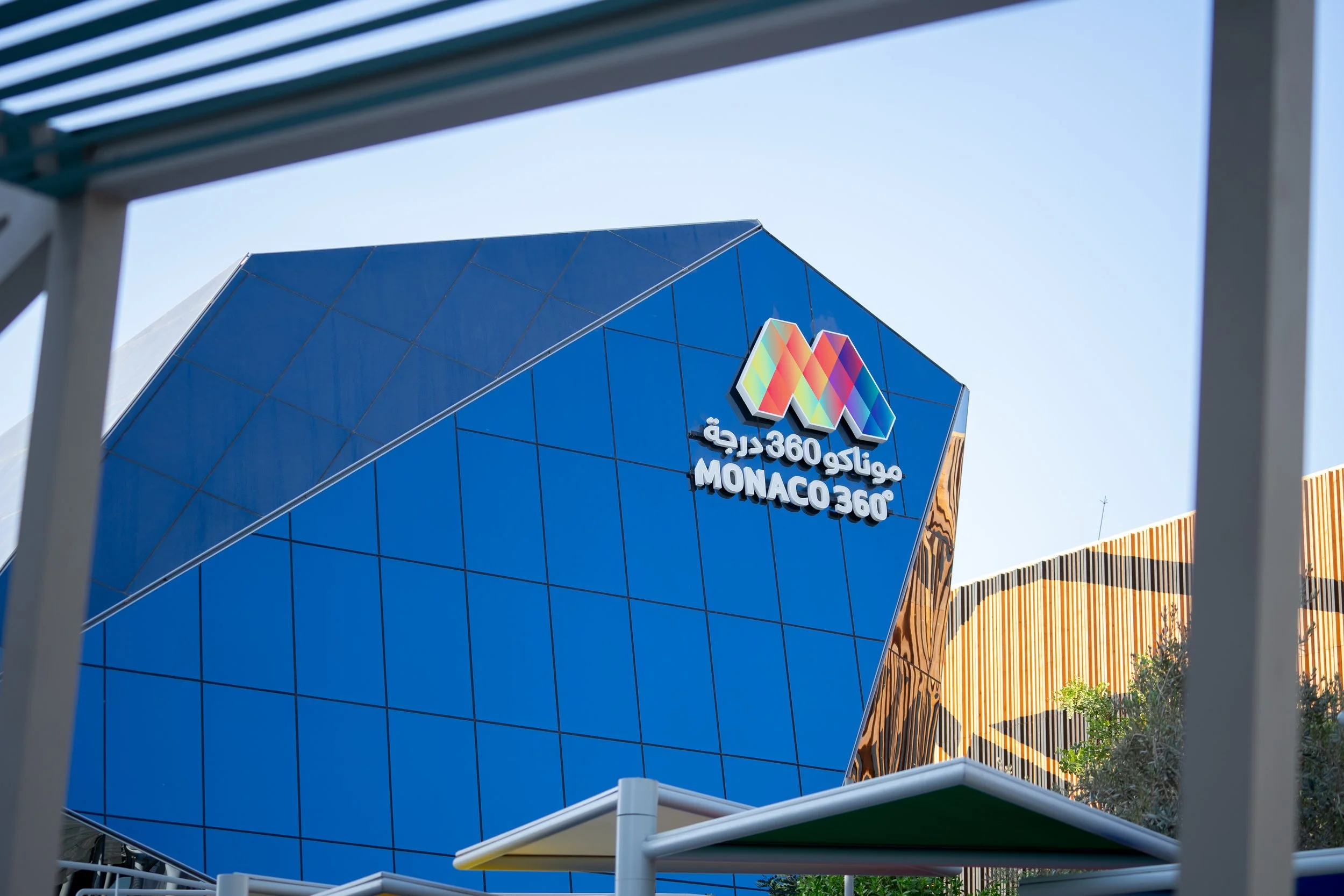 Façade d'un bâtiment moderne avec un panneau en forme de M multicolore et des textes en arabe et en français 'Monaco 360'.