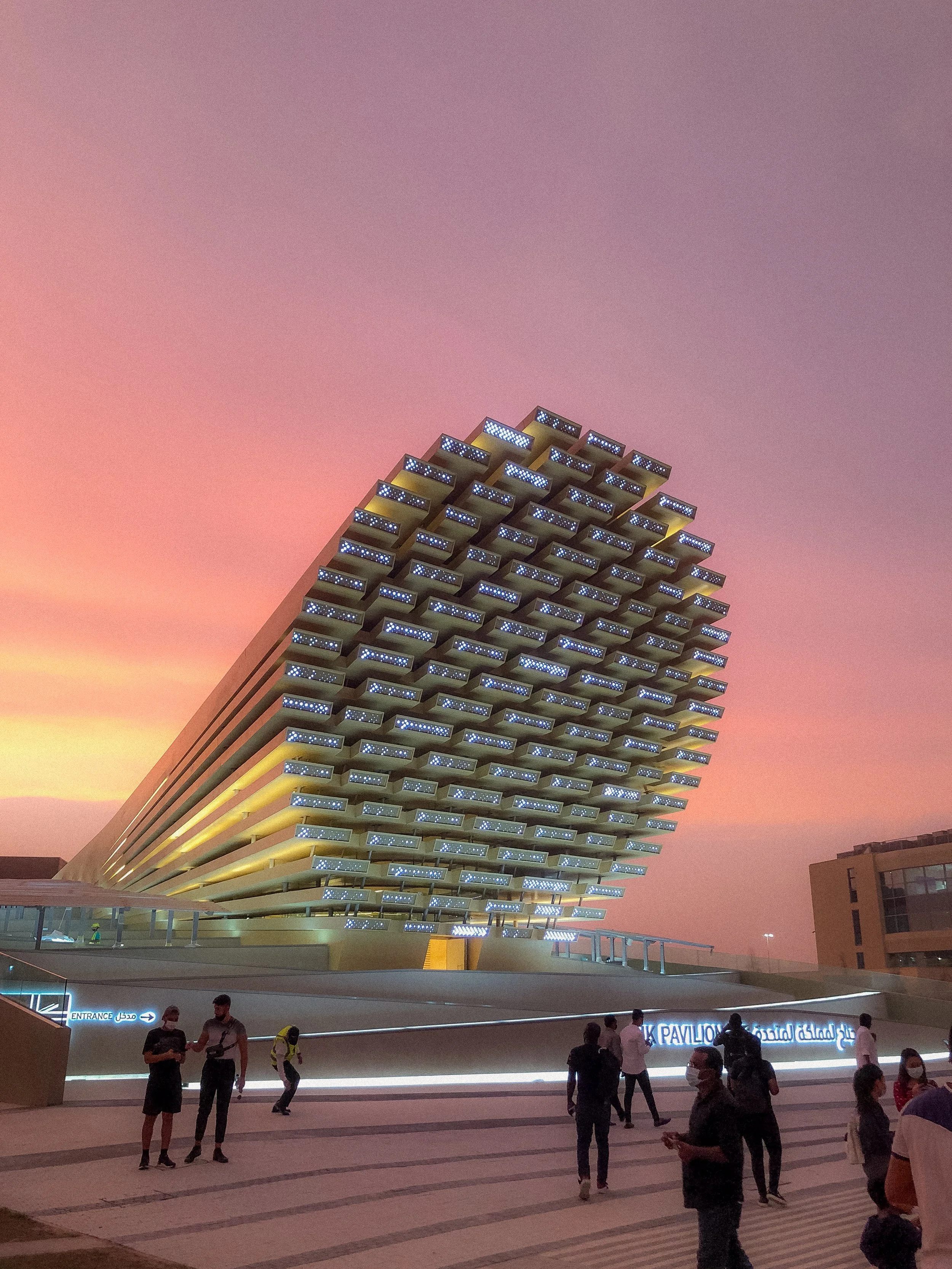 Un bâtiment futuriste en forme de voile avec des lumières bleues nocturnes, situé dans un environnement urbain au coucher du soleil.