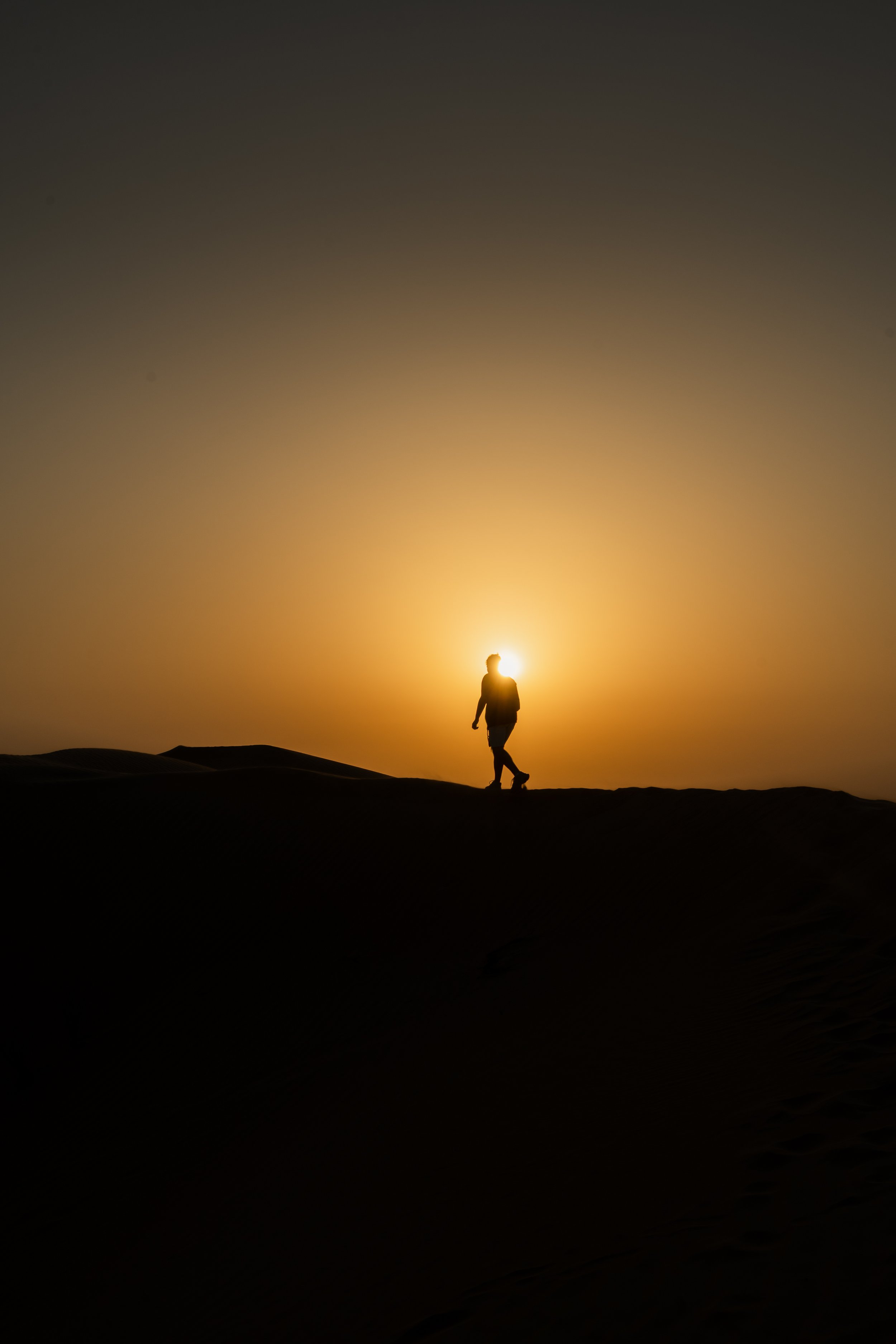 Silhouette d'une personne marchant dans un désert au coucher du soleil avec un ciel en dégradé de couleurs chaudes.