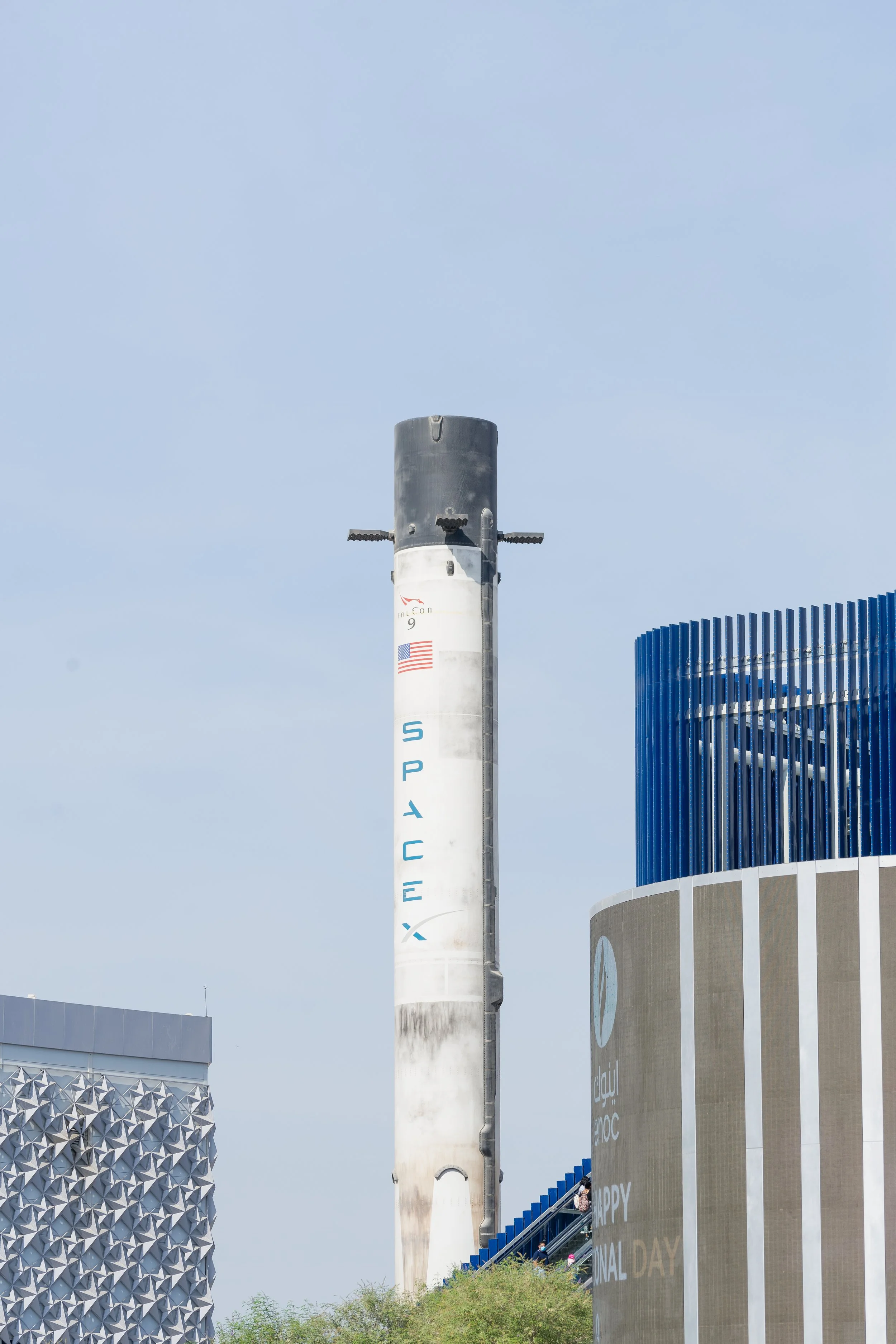 Un bâtiment futuriste avec une tour de lancement, une façade en forme de pyramides en aluminium et une partie d'un mur avec des inscriptions en anglais et en arabe, dans un environnement en plein air avec un ciel bleu clair.