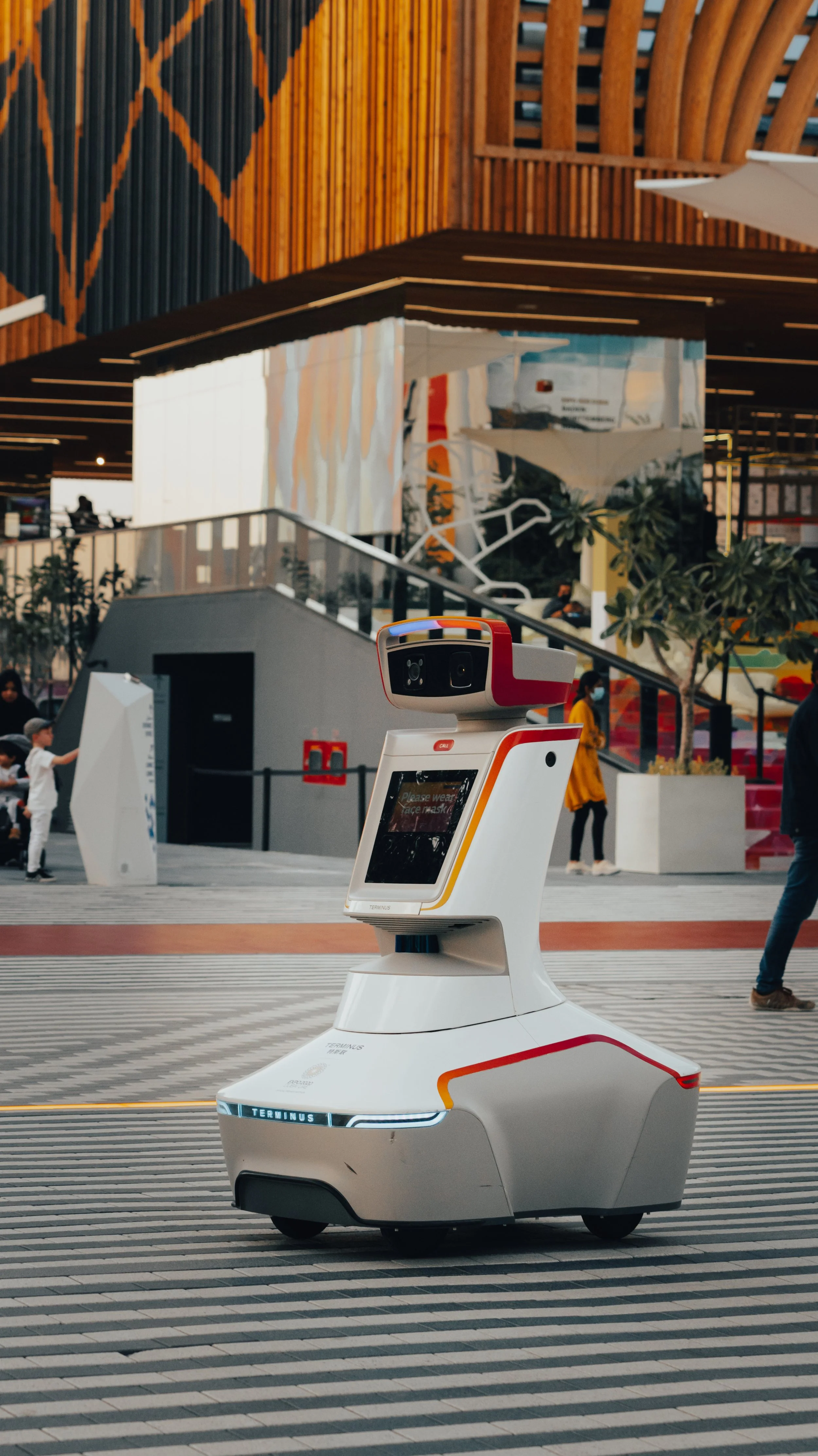 Un robot autonome dans un lieu public, probablement un aéroport ou une centre commercial, avec des personnes en arrière-plan. Le robot est blanc avec des accents rouges et jaunes, doté d'une tête avec des capteurs ou des caméras.