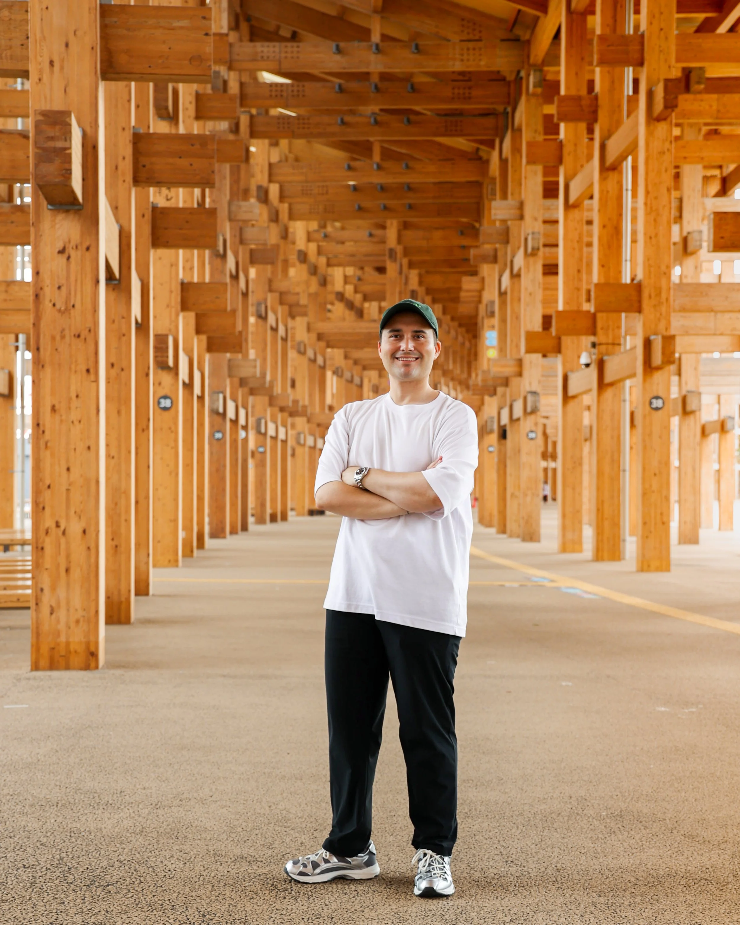 Homme souriant en tenues décontractées, debout dans un espace en bois avec une architecture à colonnes en bois, sol couvert de tapis beige, portant une casquette, un t-shirt blanc, un bracelet et des chaussures de sport.