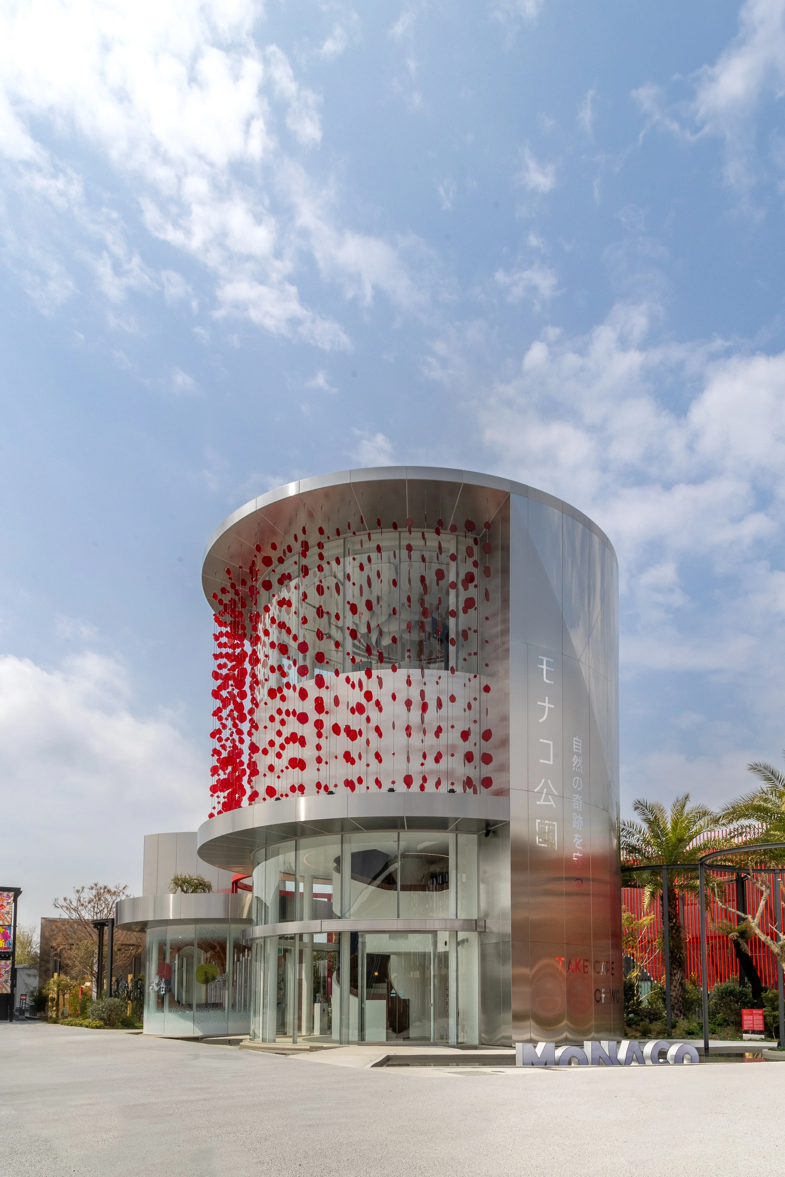 Bâtiment moderne avec façade en verre et décoration de rubans rouges, située dans un espace ouvert ensoleillé avec quelques arbres et une pancarte 'MONACO' en lettres blanches à l'avant.