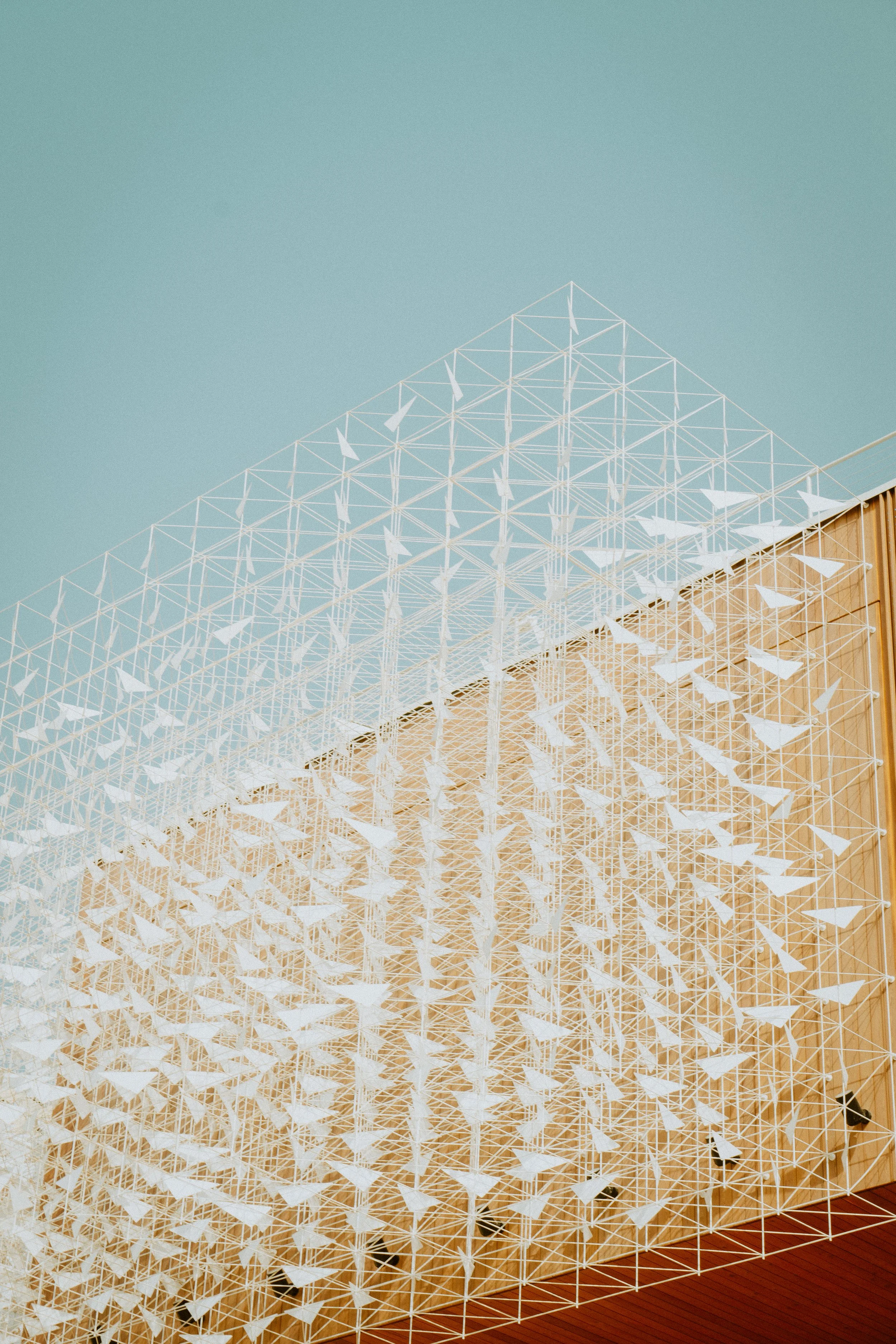 Structure géométrique composée de triangles blancs, située à l'intérieur d'un bâtiment avec un mur en bois, sous un plafond en bois, et un sol en carrelage rouge.