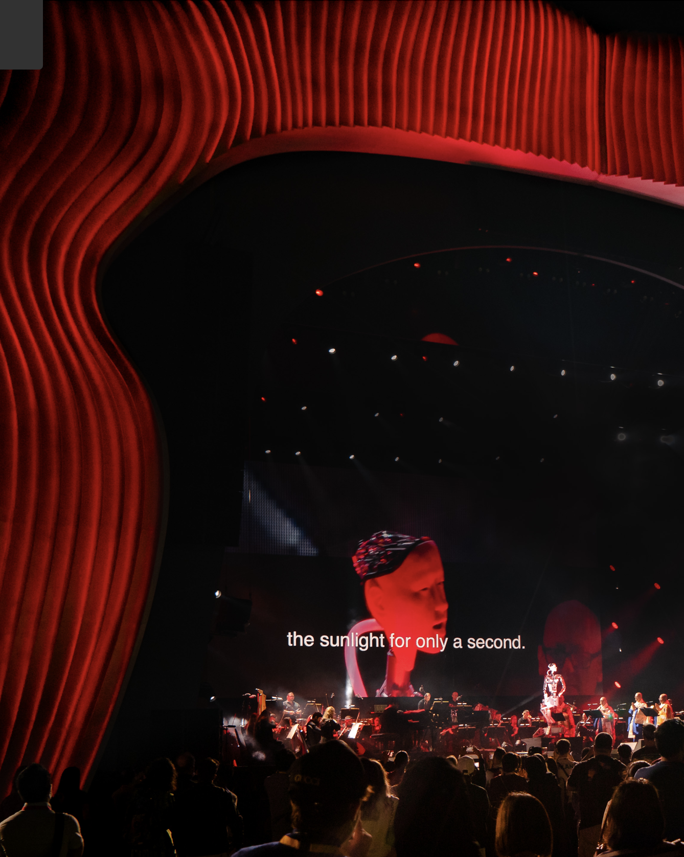 Concert avec un orchestre sur scène, lumières rouges et divers instruments, public regardant la scène