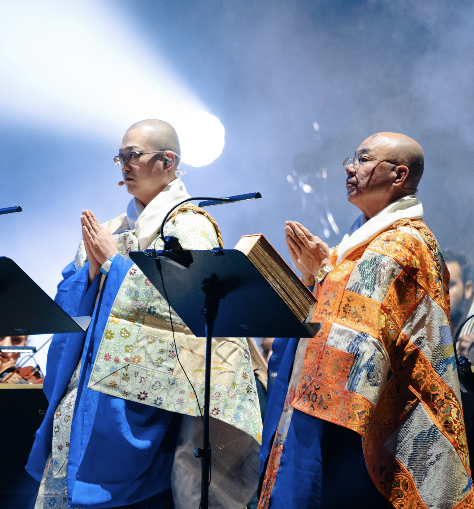 Deux hommes portant des robes traditionnelles orientales prient avec les mains jointes lors d'une cérémonie. Ils portent des lunettes et ont la tête rasée. L'arrière-plan est sombre avec une lumière intense venant de la gauche.