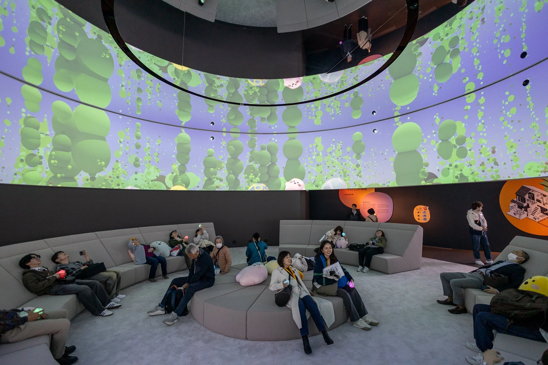 Une salle d'attente dans un musée ou un centre scientifique avec des visiteurs assis sur des bancs, regardant une grande projection lumineuse au plafond représentant des bulles vertes contre un fond violet.