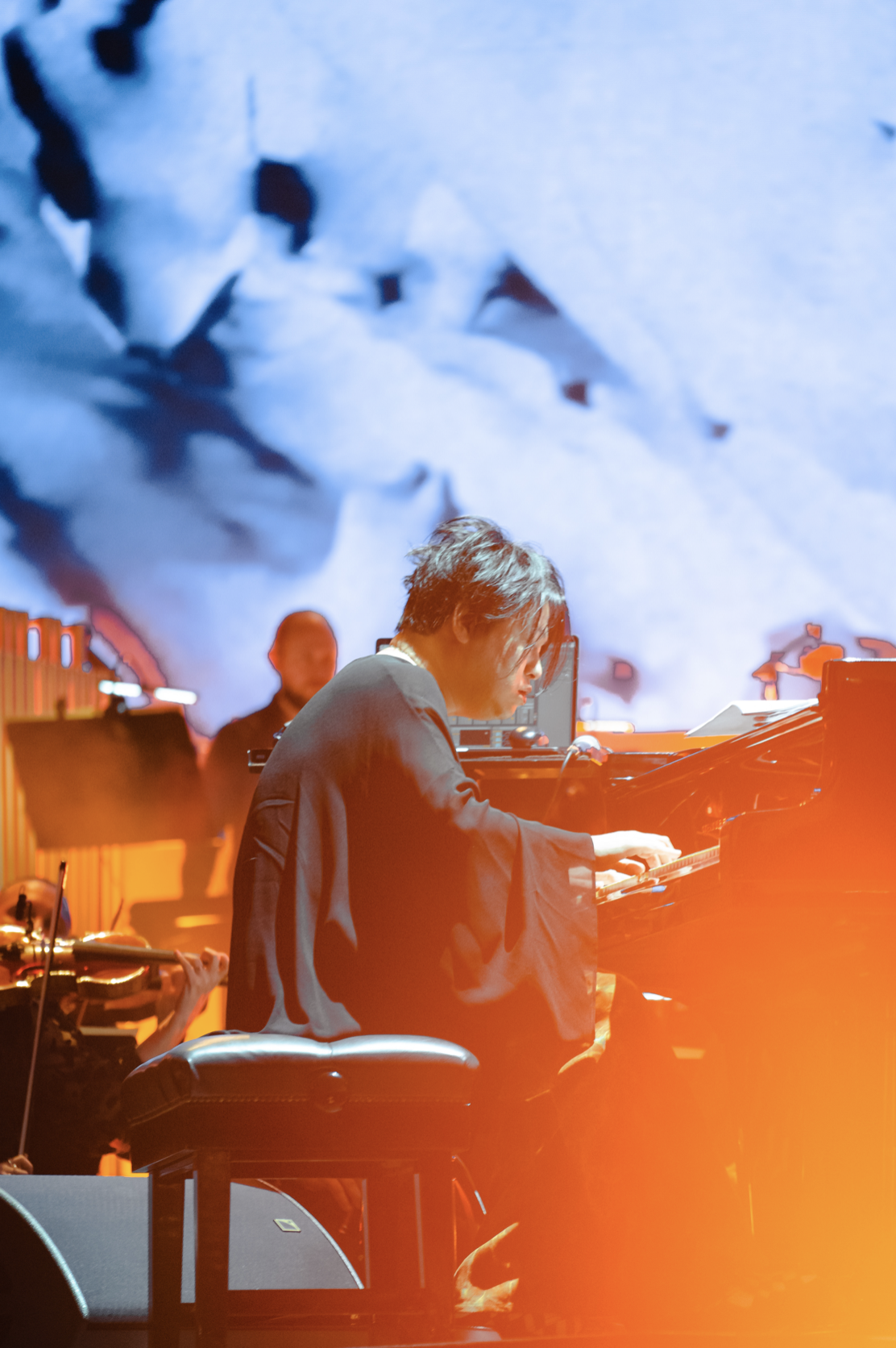 Un musicien joue du piano sur scène avec un fond de projection abstraite en bleu et rouge.