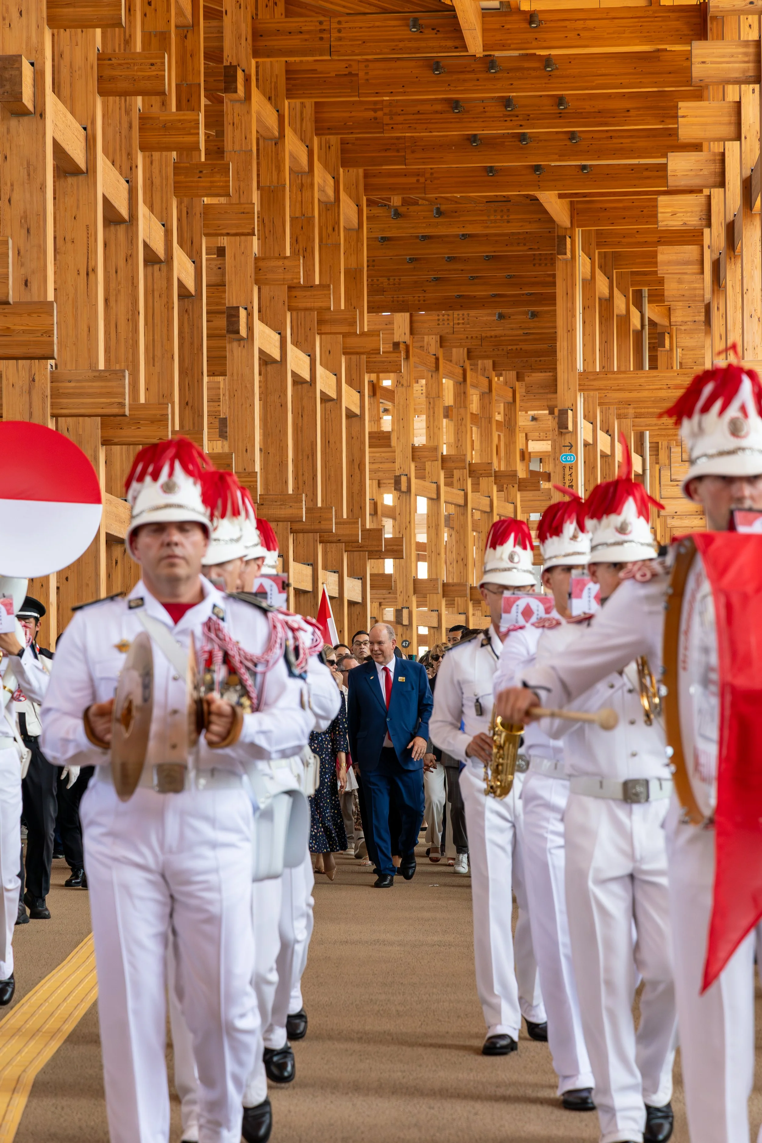 Groupe de personnes en uniforme blanc, portant des chapeaux avec des pompons rouges, marchant dans une procession avec une structure en bois en arrière-plan.