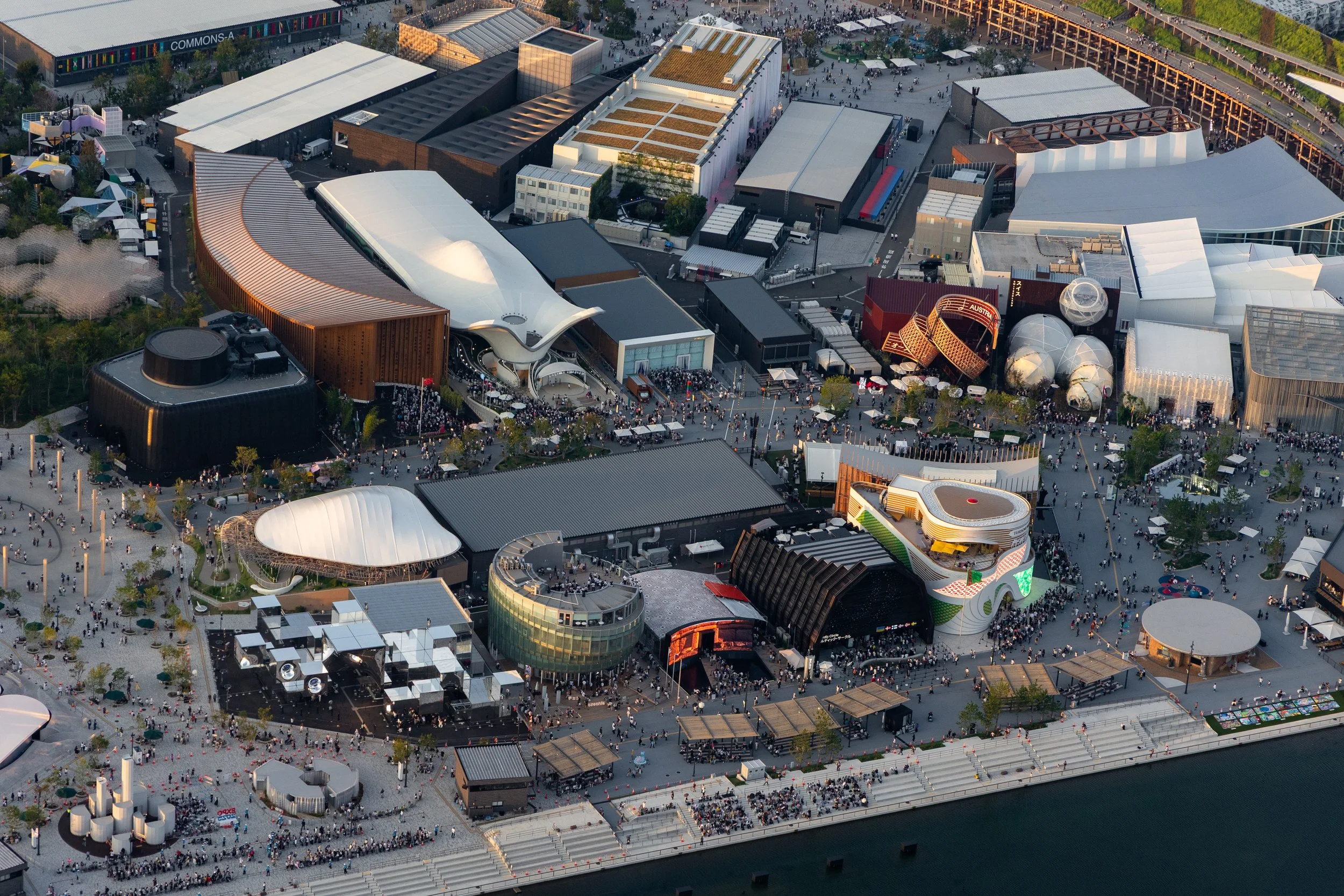Une vue aérienne d'un grand festival ou événement avec de nombreux bâtiments modernes, un grand nombre de visiteurs, des espaces d'exposition et des zones de divertissement en plein air dans une zone urbaine près de l'eau.