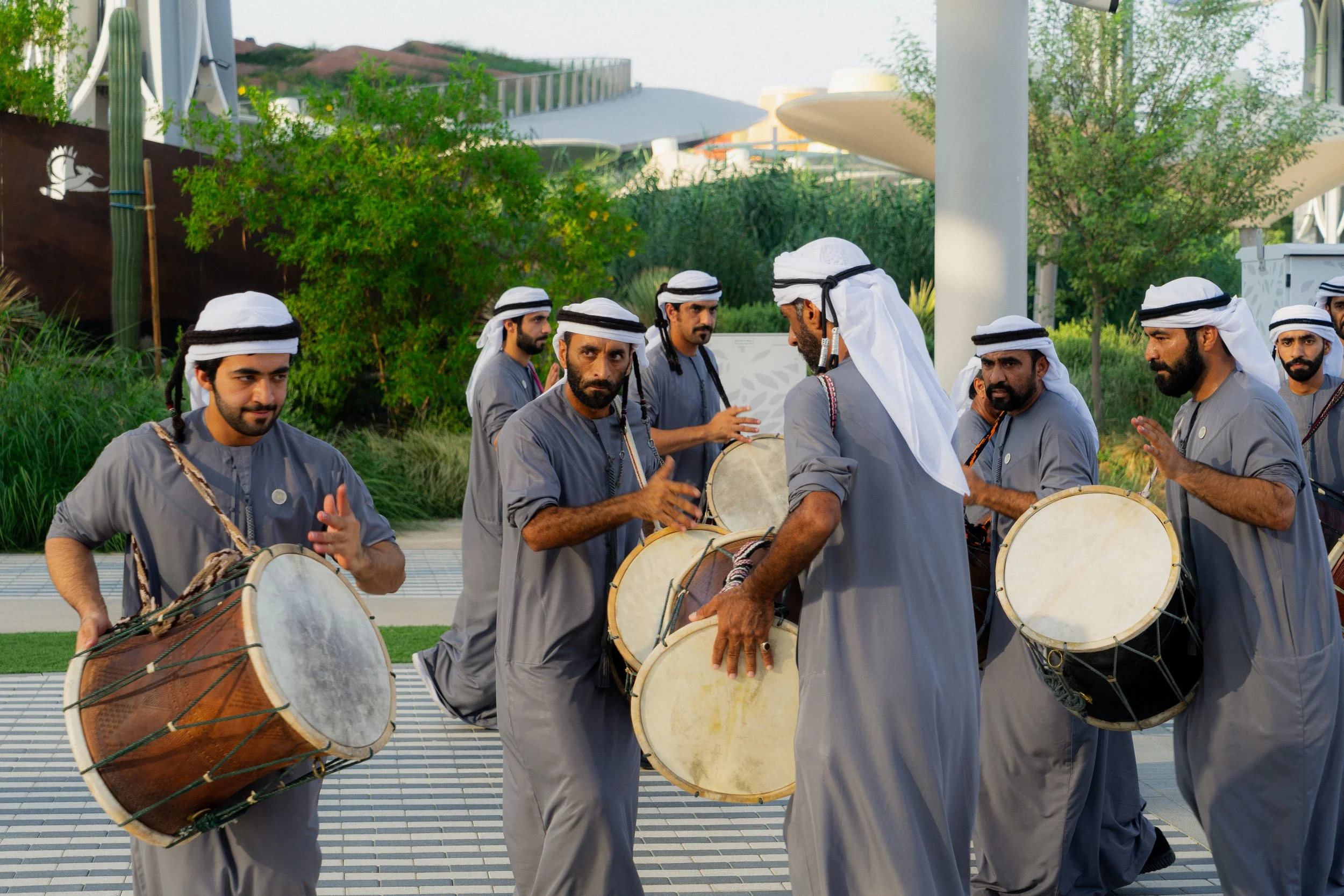 Groupe d'hommes en vêtements traditionnels jouant du tambour lors d'une performance extérieure, avec des arbres et une structure moderne en arrière-plan.