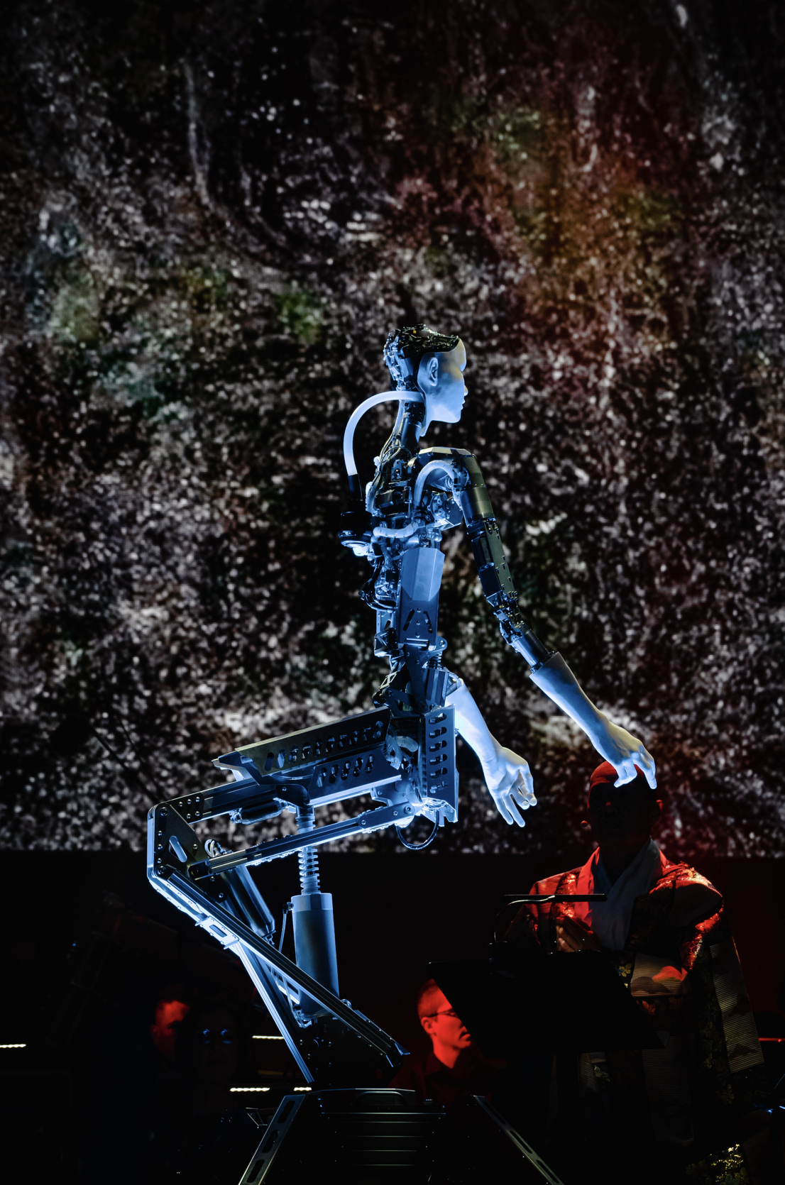 Un robot humanoïde est debout, touchant une personne assise. Le robot a une apparence métallique et est éclairé en bleu. En arrière-plan, un écran affiche des motifs lumineux.