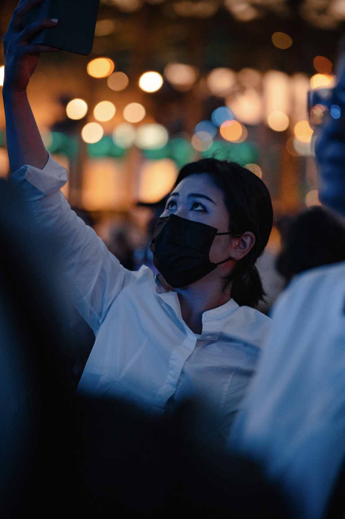Une femme portant un masque noir, portant une chemise blanche, semble prendre une photo ou un selfie lors d'un événement nocturne.