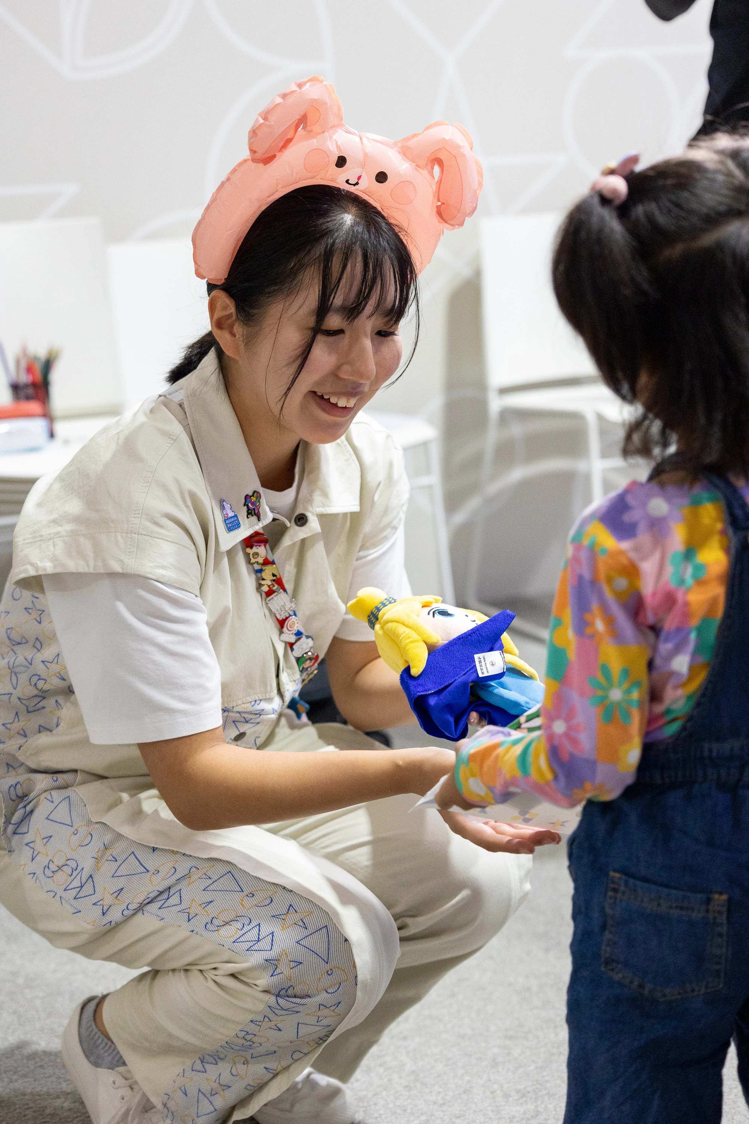 Une femme en uniforme de soignante ou bénévole souriante offre un cadeau à une enfant portant un vêtement coloré. La femme porte un chapeau en forme de lapin en papier, et la scène se déroule dans un environnement intérieur avec des chaises blanches.