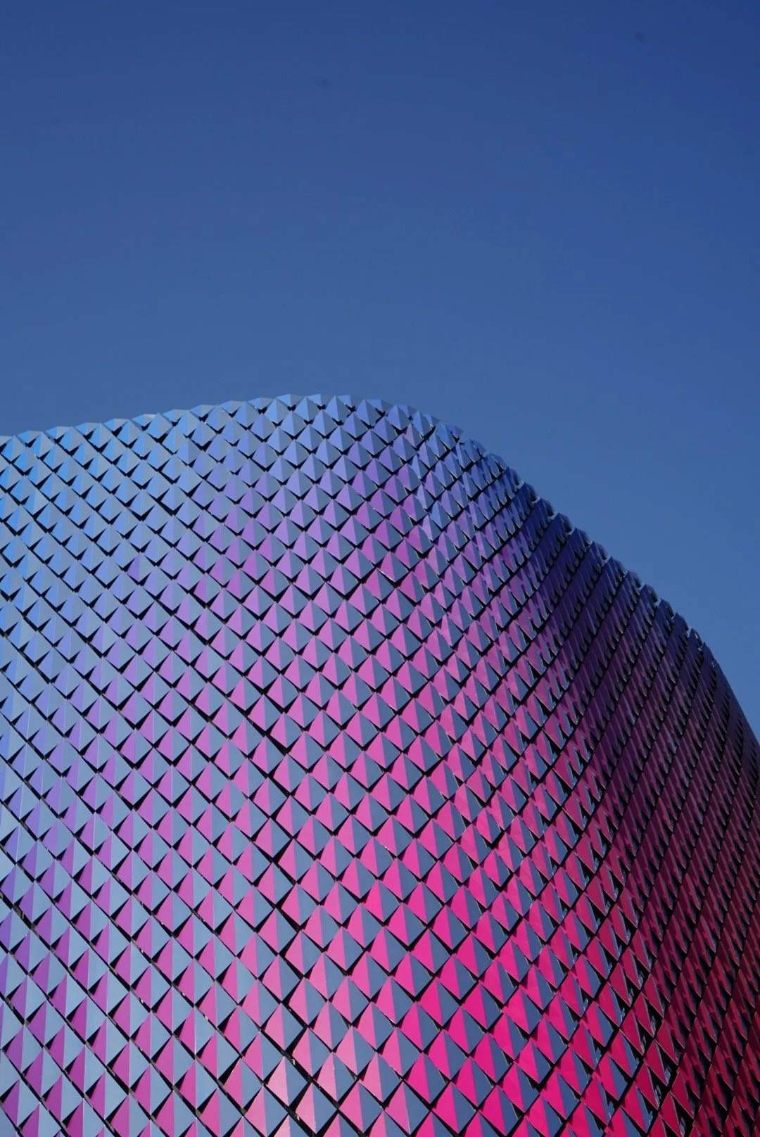 Une grande structure métallique en forme de dôme avec une surface recouverte de petits triangles colorés en violet et bleu, contre un ciel clair.