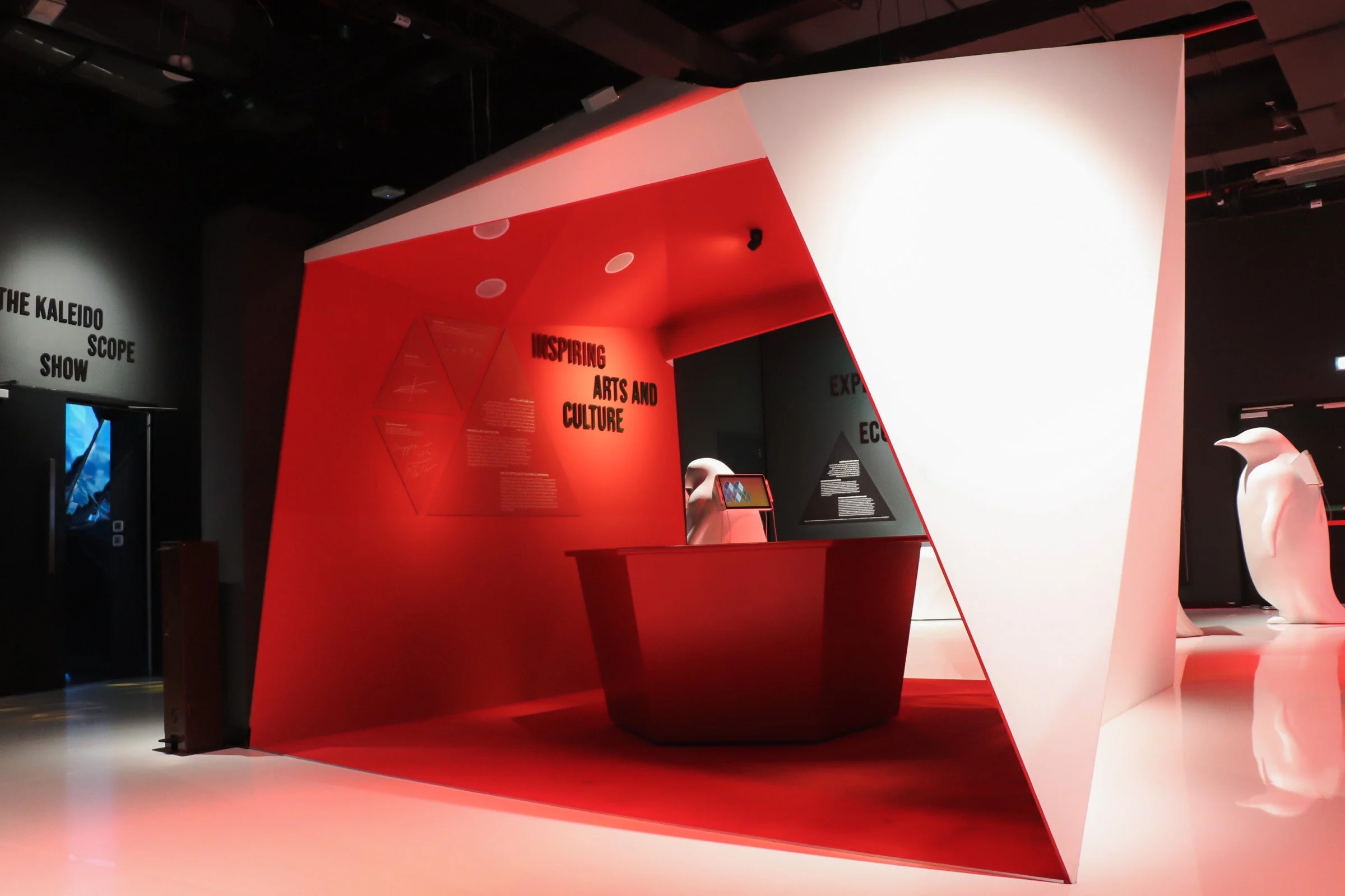 Un espace d'exposition avec un mur rouge portant le texte 'Inspiring Arts and Culture' et une structure blanche artistique. Il y a des écrans et des manchots en sculpture dans l'espace.