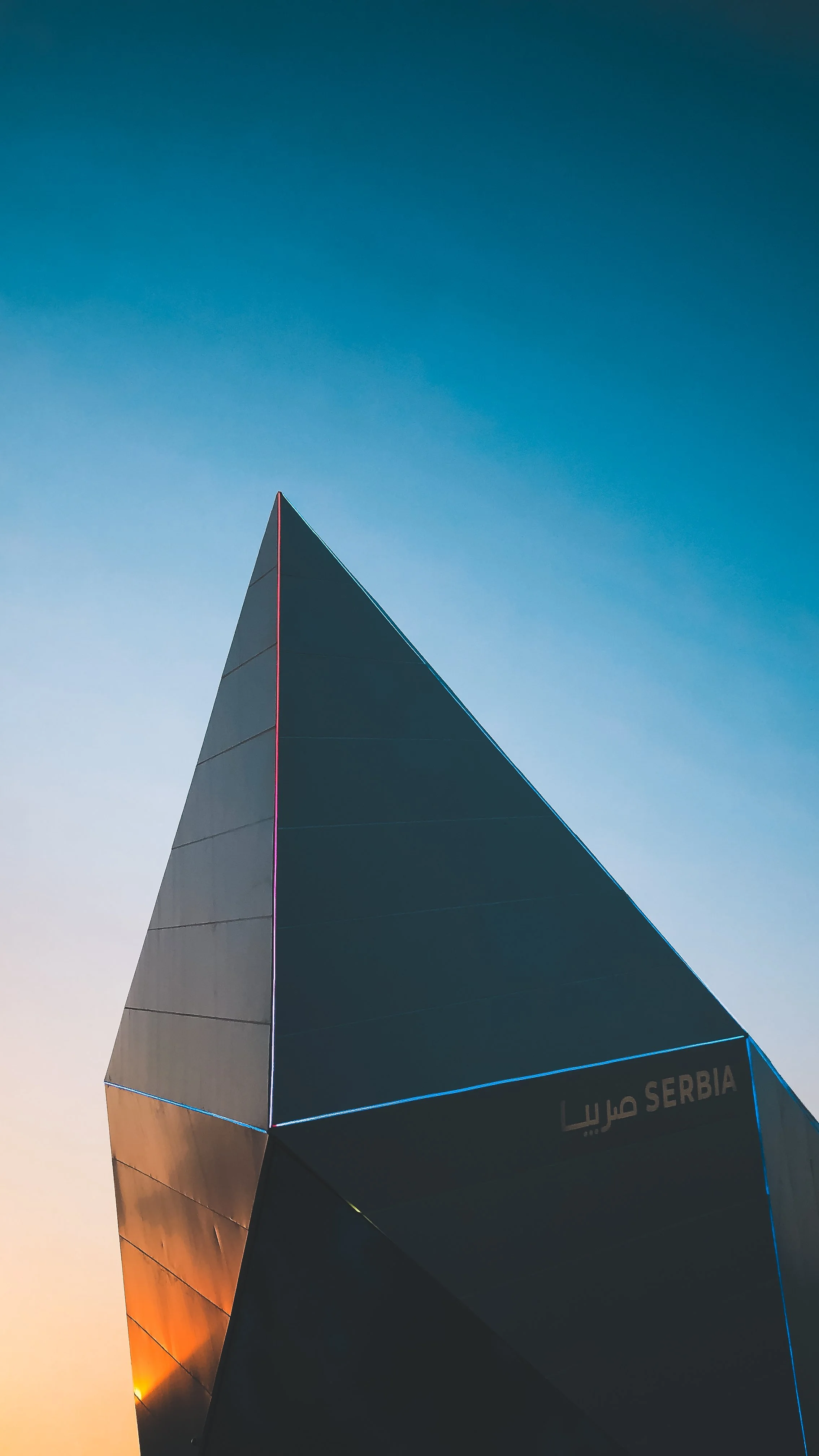 Vue d'une structure architecturale en forme de voile ou de pyramide moderne avec un ciel clair en arrière-plan, la structure porte l'inscription 'Serbia' en anglais et en arabe.