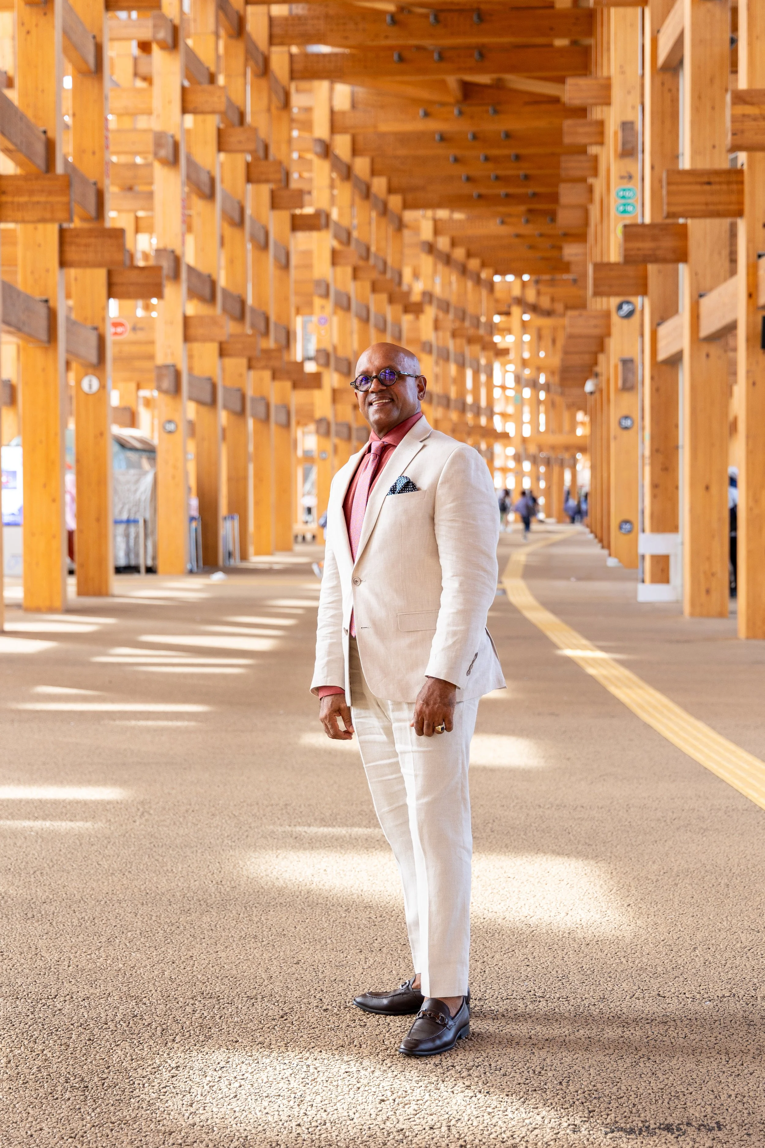 Un homme en costume beige, chemise rose, cravate rose, lunettes rondes, souriant, dans un bâtiment en bois avec structure en bois visible, sol en moquette, quelques personnes en arrière-plan.
