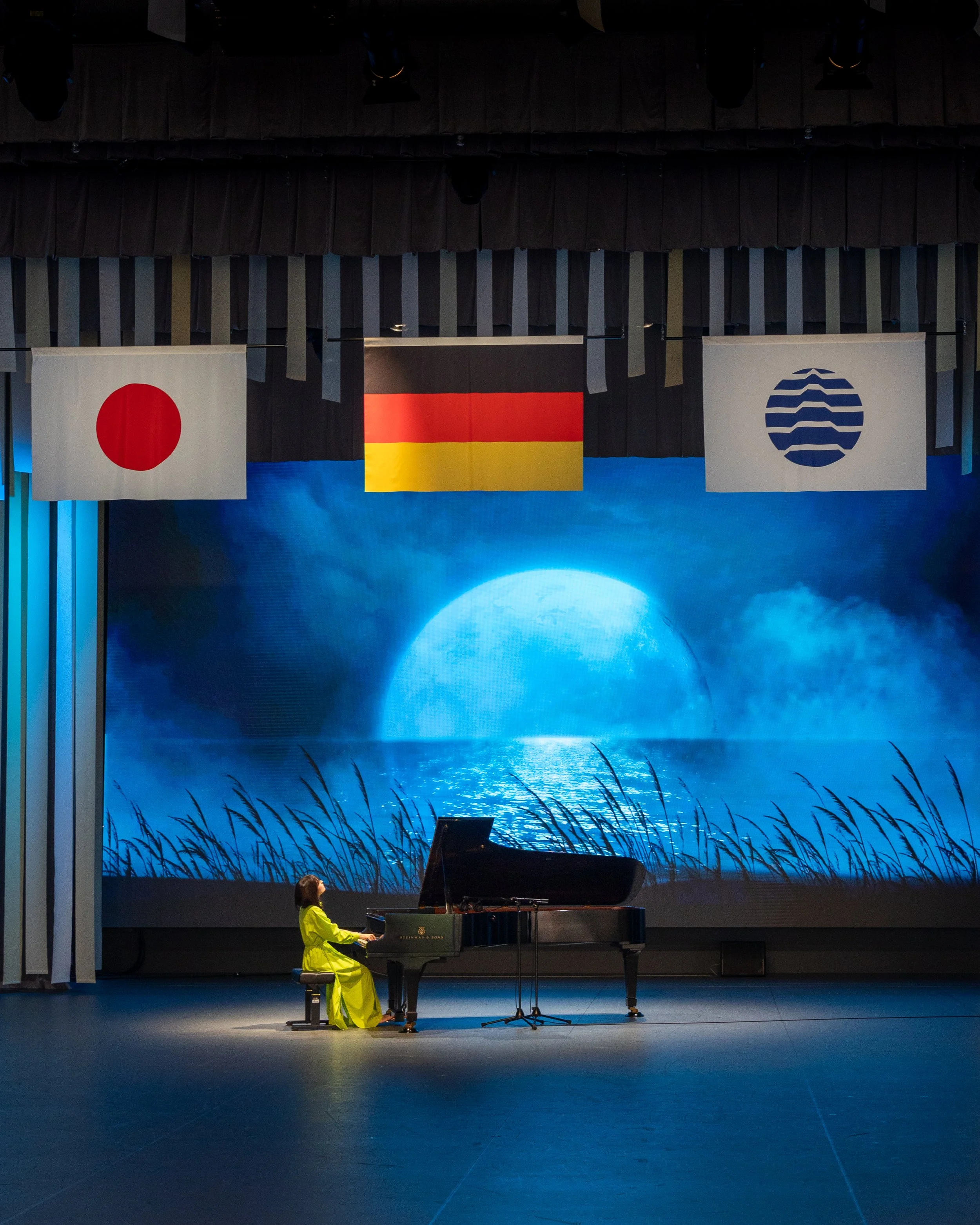Une personne joue du piano sur scène, portant une tenue jaune vif, dans une salle de concert avec un décor représentant un lac ou une étendue d'eau et des montagnes en arrière-plan, avec des drapeaux de Japon, Allemagne et pays sans nom visible suspe