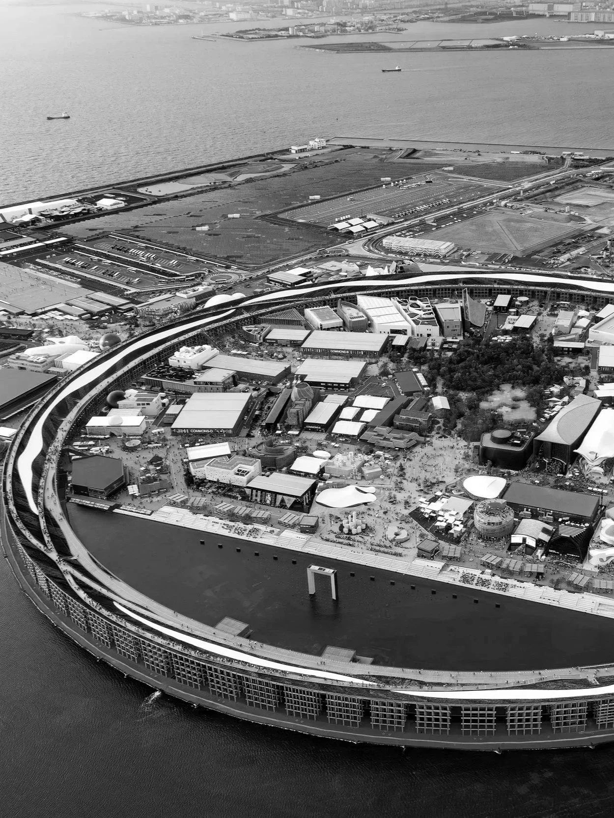 Vue aérienne d'une ville avec un bâtiment en forme de cercle près de l'eau, entouré de structures modernes et de zones vertes.