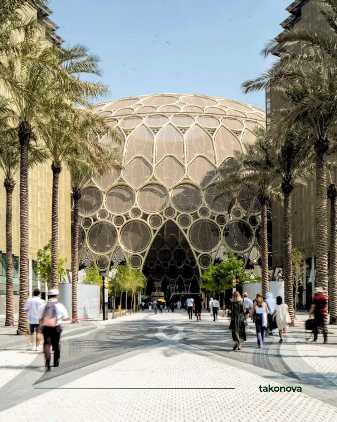 Un bâtiment futuriste avec une façade en forme de cercle et de motifs géométriques, entouré de palmiers, avec des personnes marchant devant dans une zone piétonne.