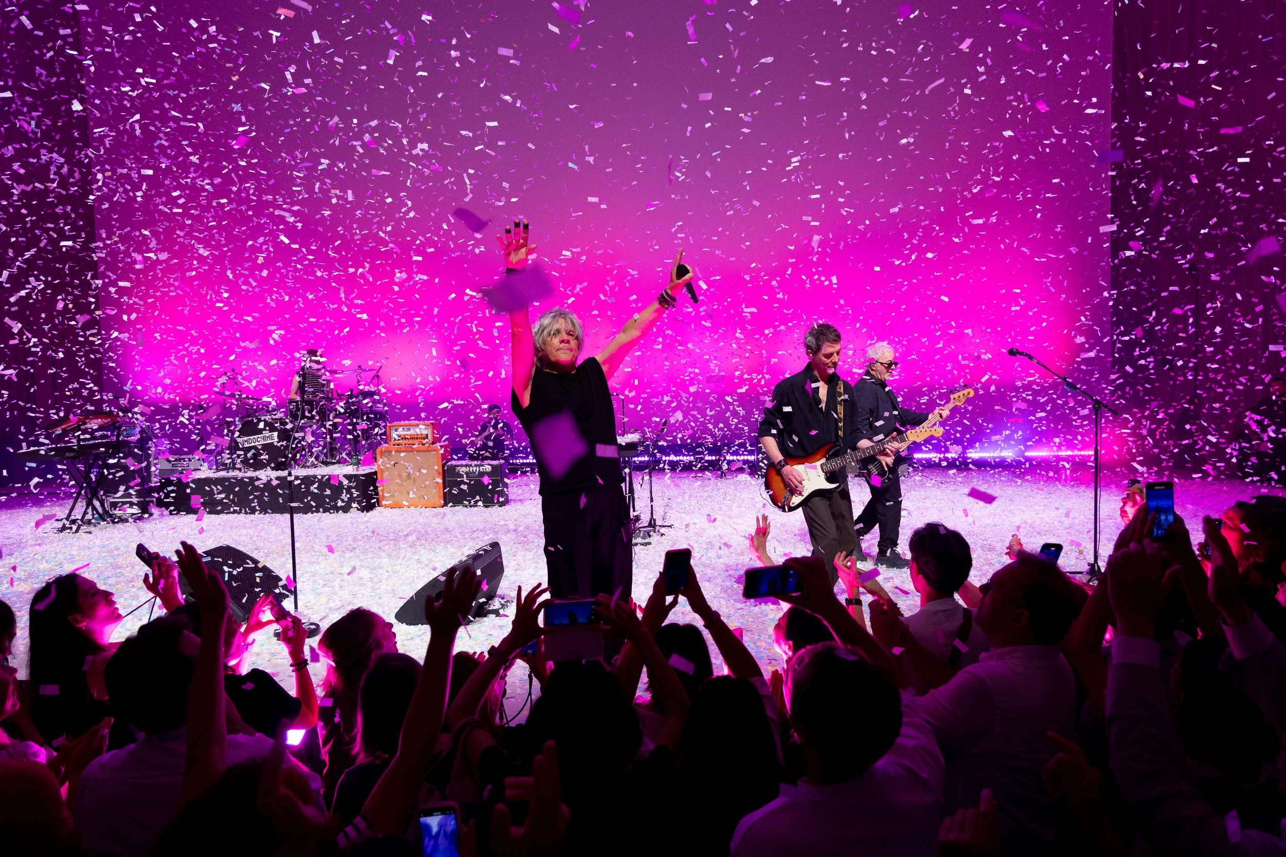Un groupe de musiciens sur scène en train de jouer, entourés de confettis rose et blancs, avec des spectateurs en première ligne et des lumières violettes en fond.