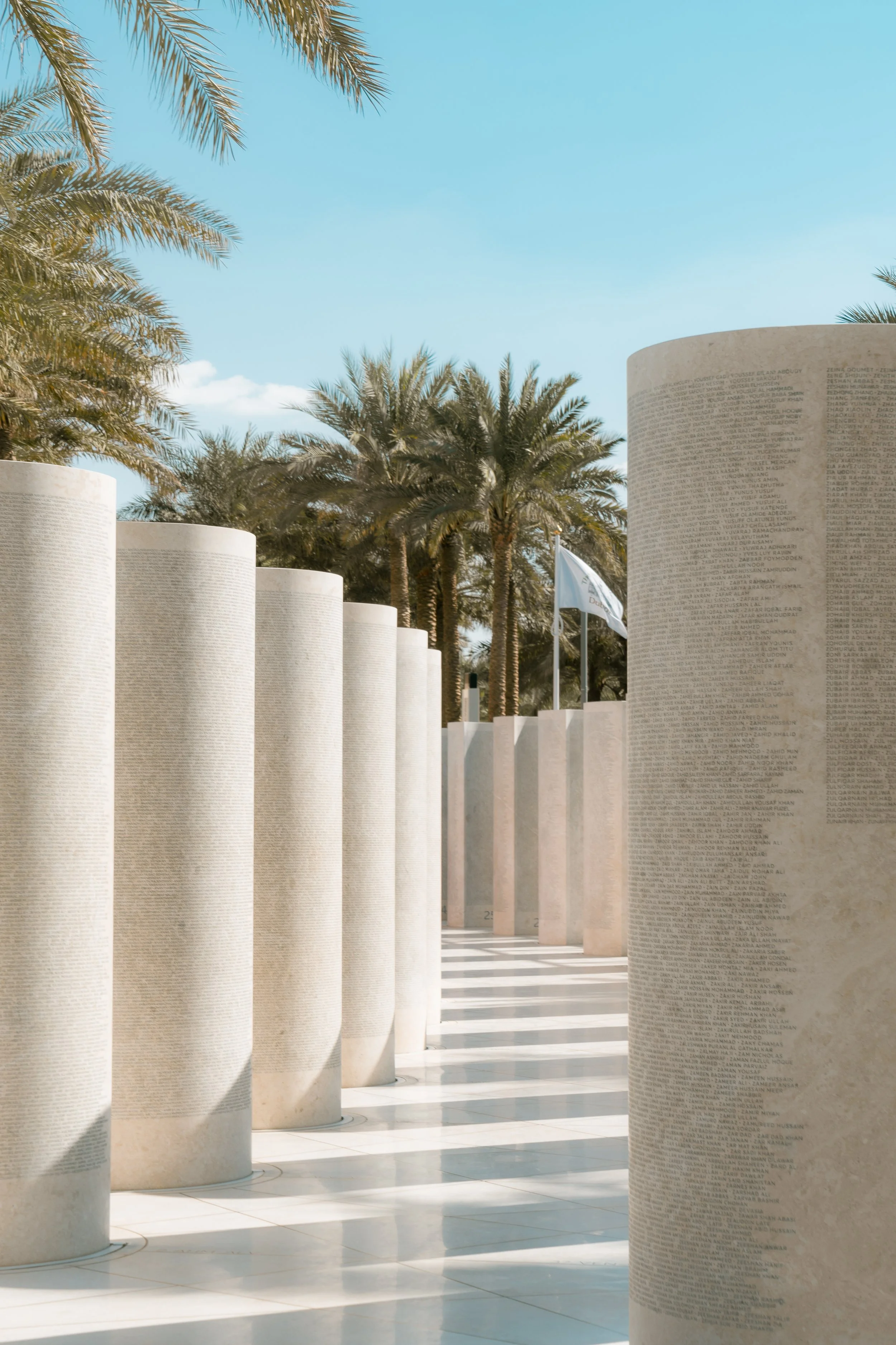 Murs cylindriques en pierre gravés avec des noms, entourant une zone avec des palmiers et un ciel bleu clair, symbole de mémoire ou d'hommage.