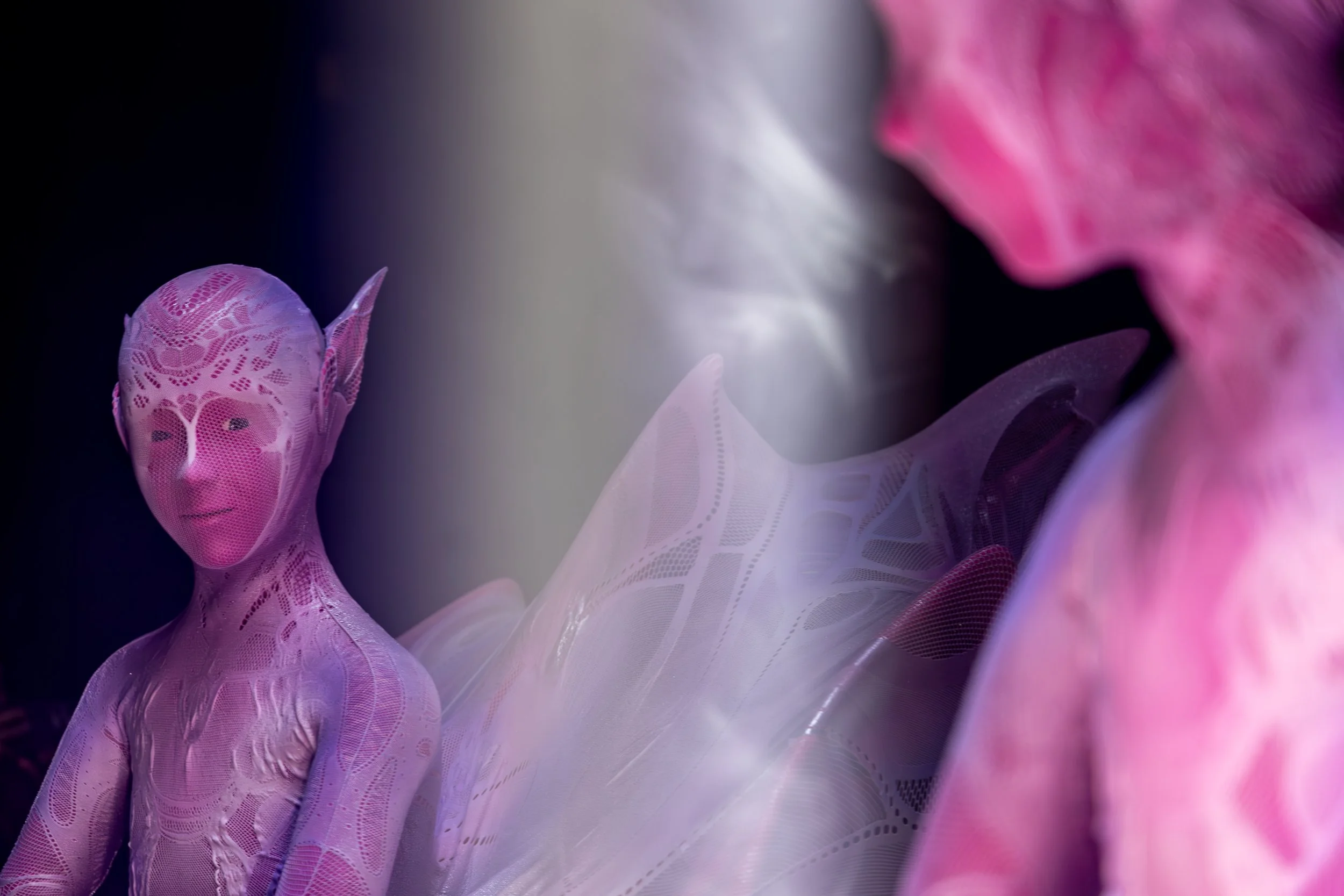 Une sculpture ou une création artistique représentant une figure humaine avec une peau en tissu délicat et translucide, principalement en nuances de rose et violet, entourée de formes abstraites.